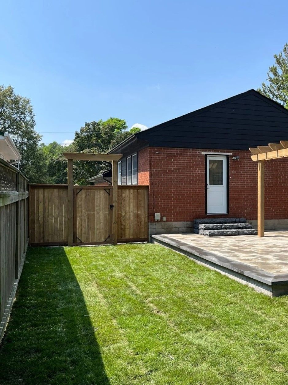 A backyard view with a wooden fence and gate, a brick building with a white door, and a paved patio area.