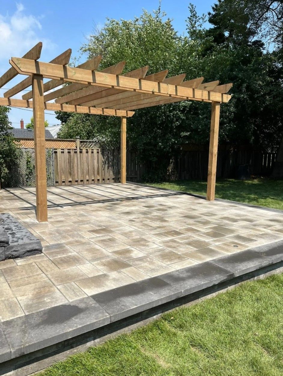 Wooden pergola over a stone patio in a grassy backyard with a dark stone border.