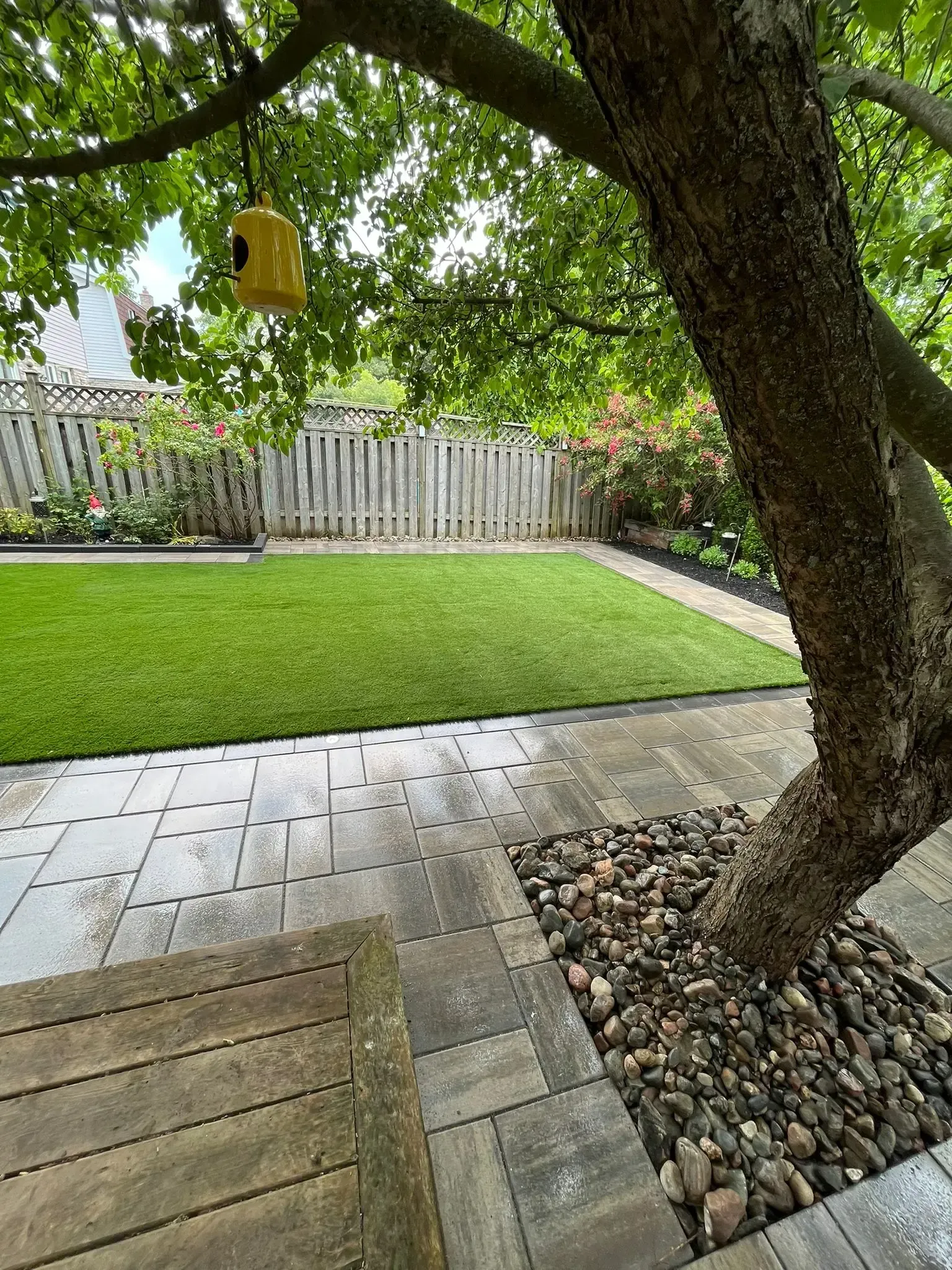 A backyard with stone patio, green lawn, tree with bird feeder, and wooden fence.