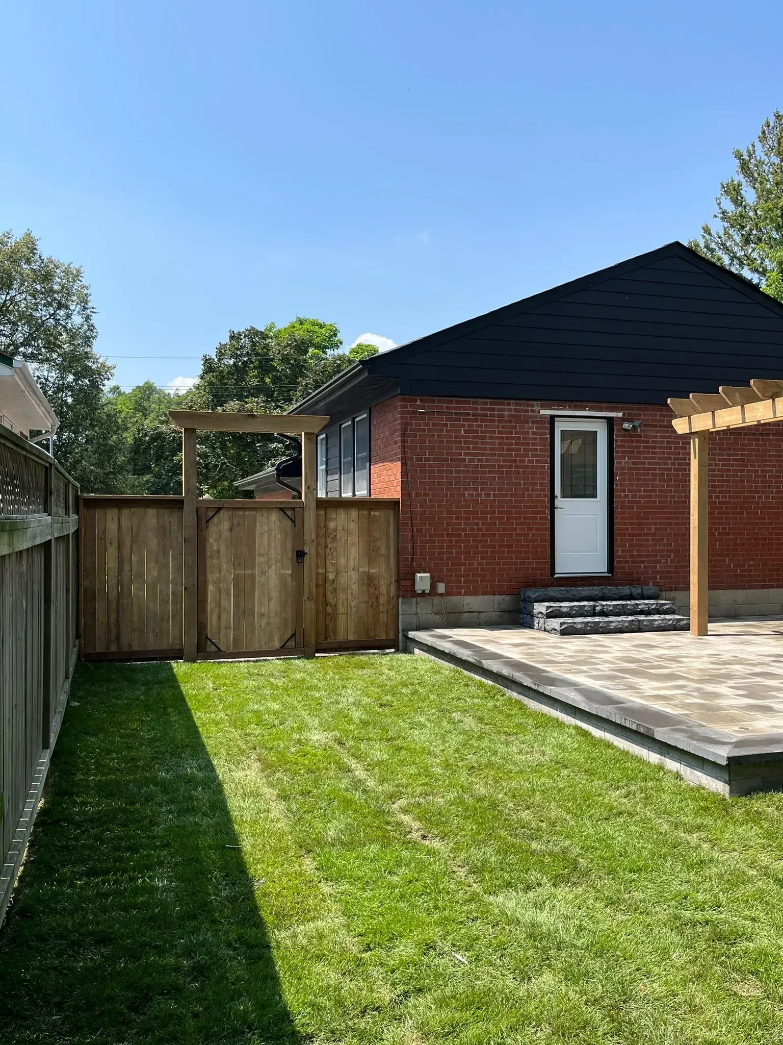 Backyard with a brick house, wooden fence, grass, and patio on a sunny day.
