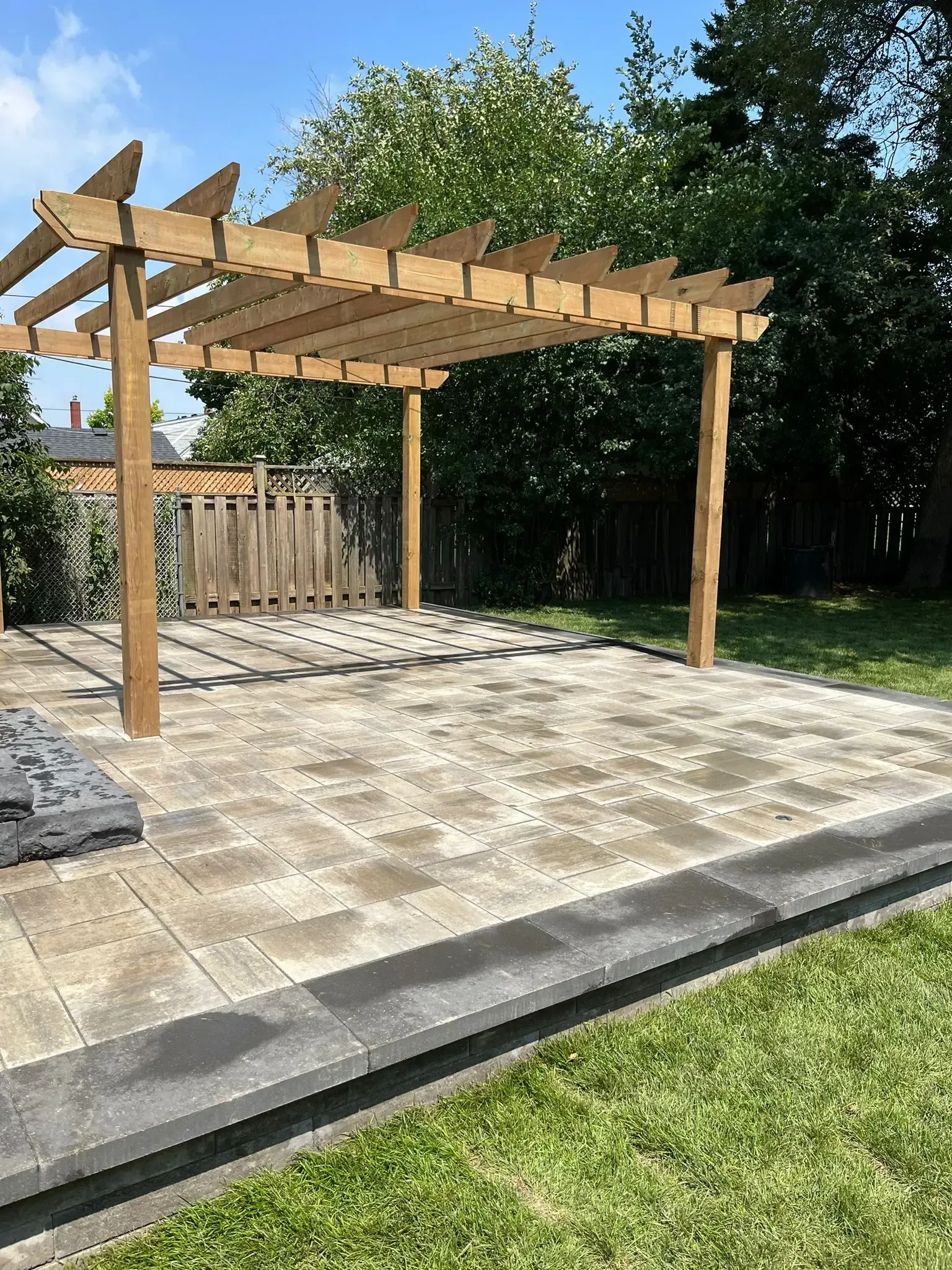 Wooden pergola over a paved patio. Green grass surrounds the patio. Blue sky in the background.