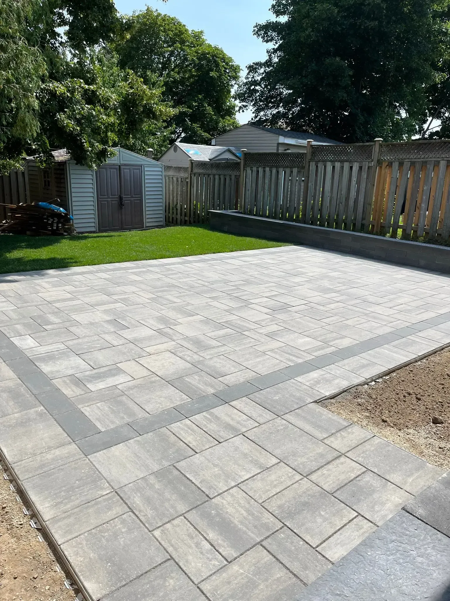 Paver patio with a dark border, bordered by grass, fence, and shed, in sunny backyard.