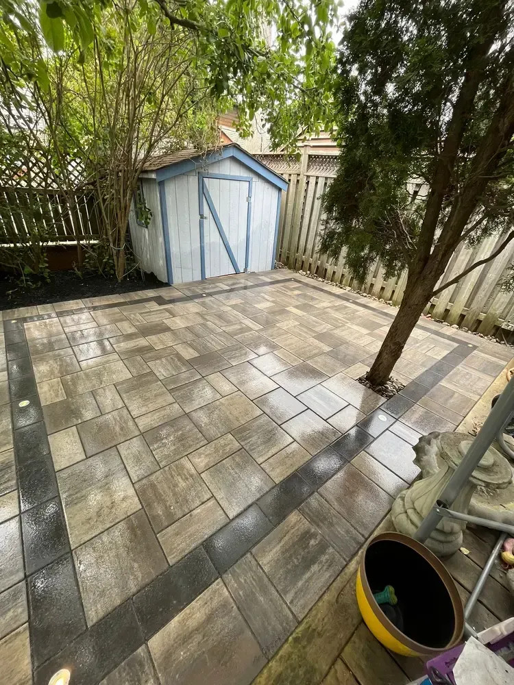 Patio with tan and black pavers, a small shed, and a tree, enclosed by a fence.