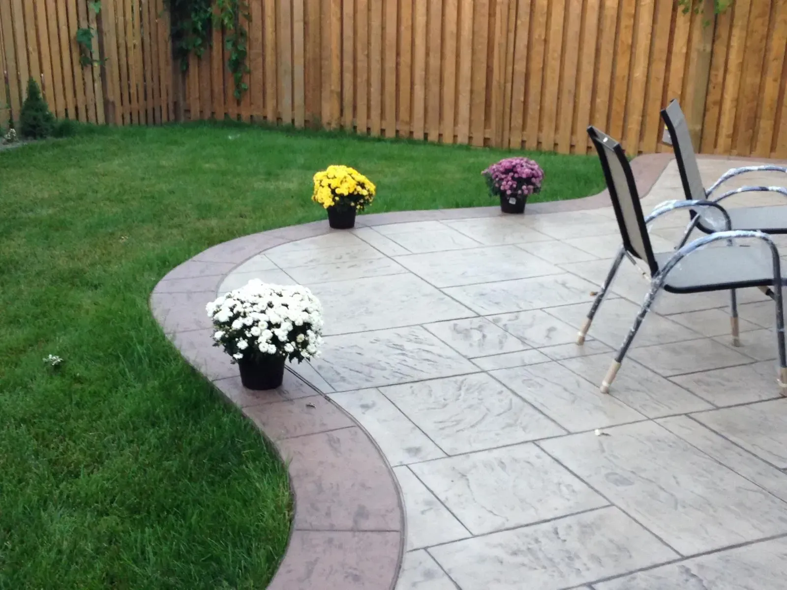 Patio with flower pots on paved surface, green lawn, wooden fence, and chairs.