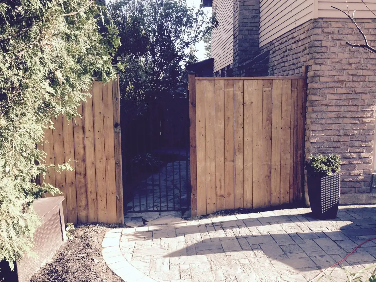 Wooden gate open to a garden, path with brick pattern, house on the right, and a potted plant.