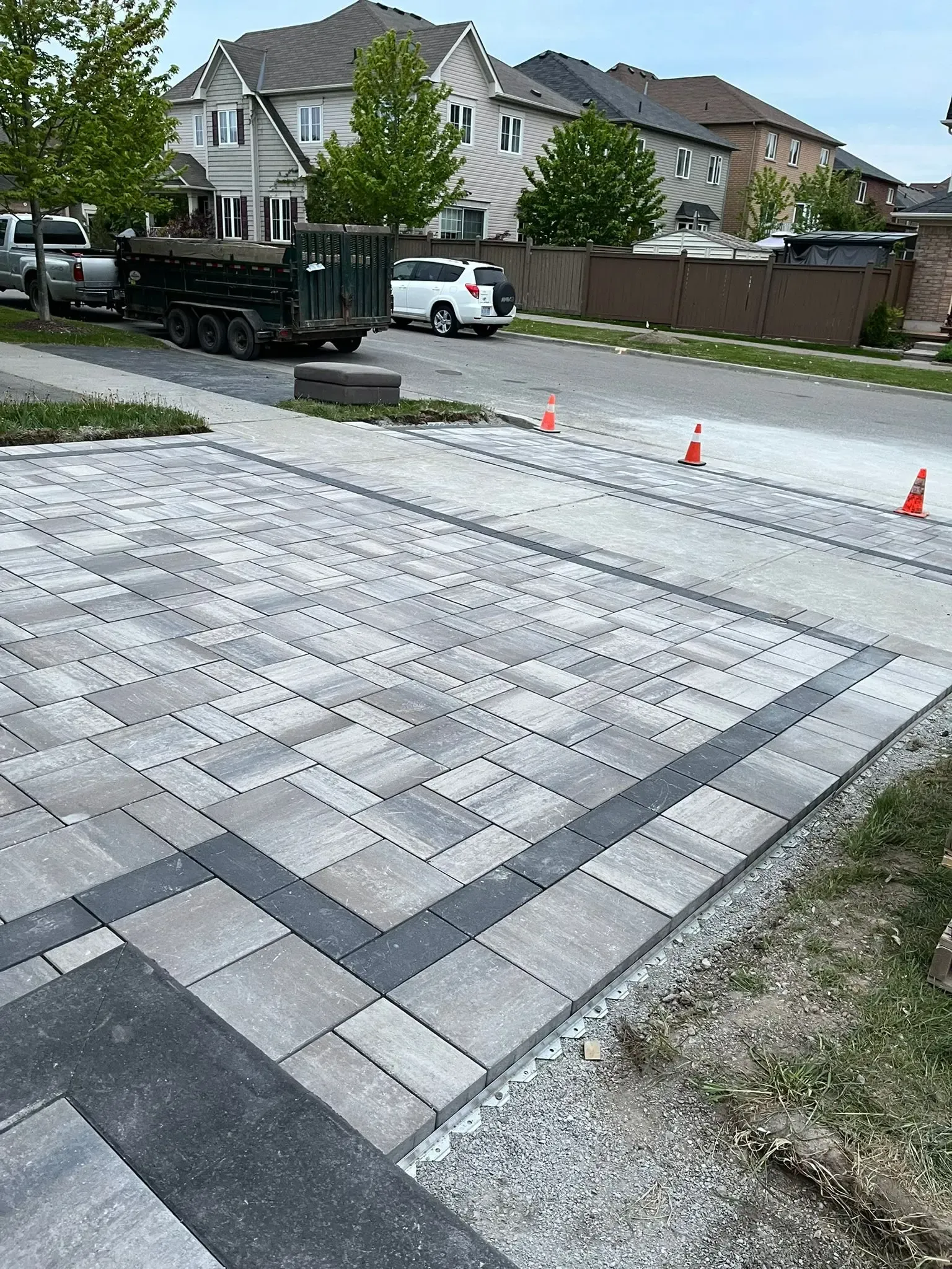 Driveway with new pavers in varying shades of gray, dark border, construction equipment in background.