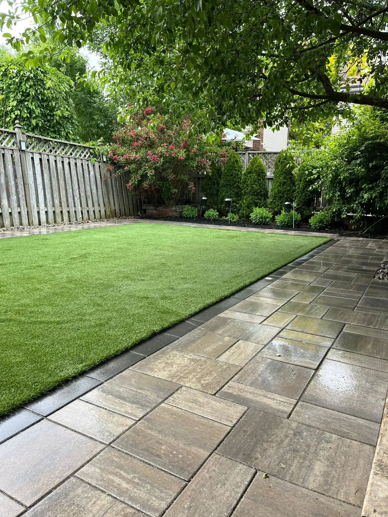 Backyard patio with paved stones and green turf, bordered by a black strip, with a fence and trees.
