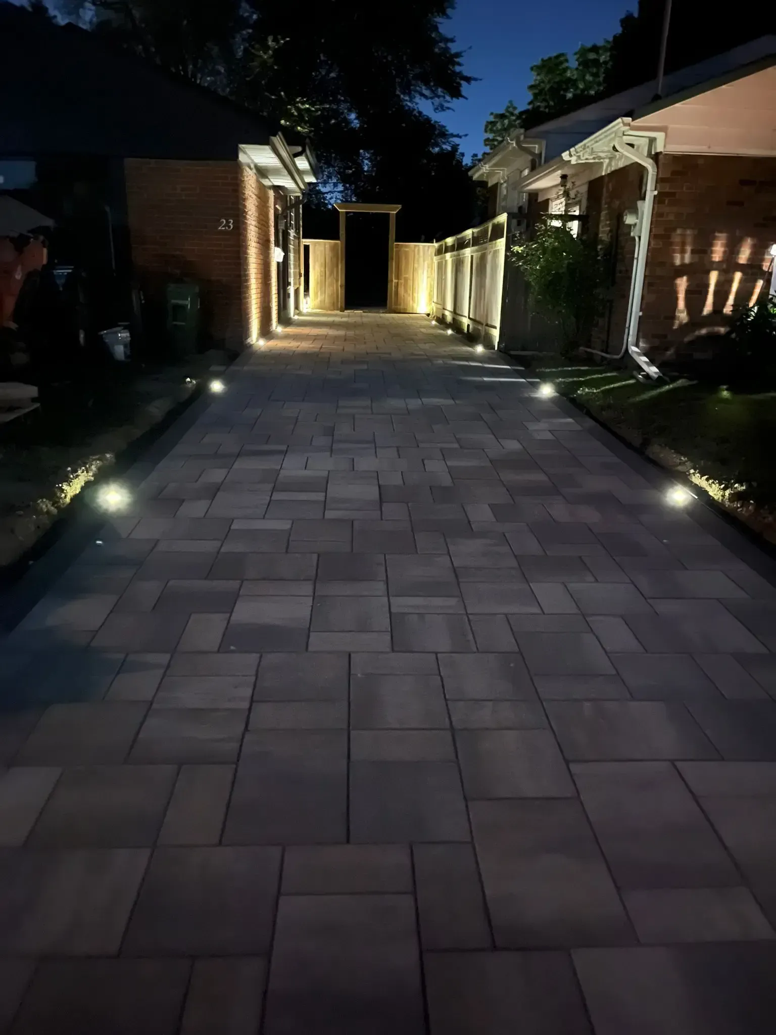 Nighttime view of a brick driveway with illuminated landscaping. Lights on the ground and buildings.