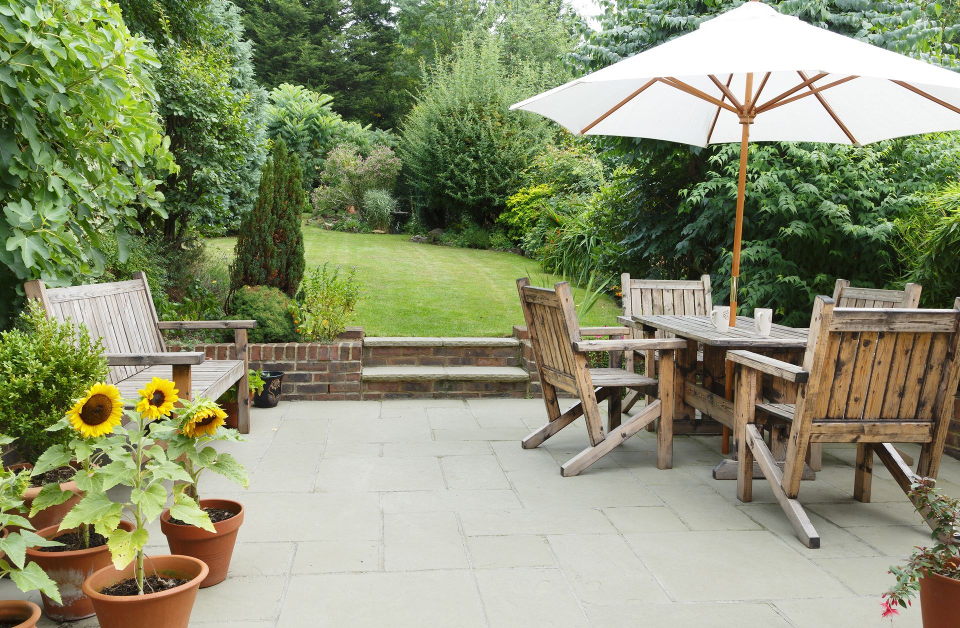 Patio with wooden furniture, umbrella, sunflowers, and lush green garden in the background.
