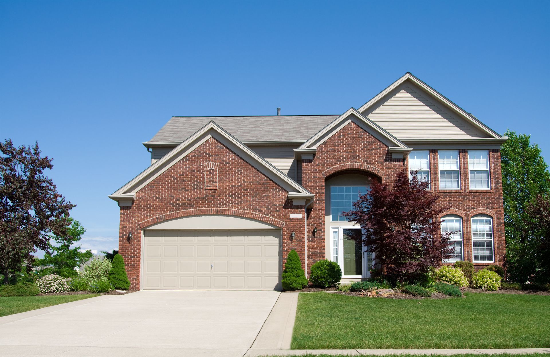 Two-story brick house with a tan garage door, set against a blue sky, with a green lawn.