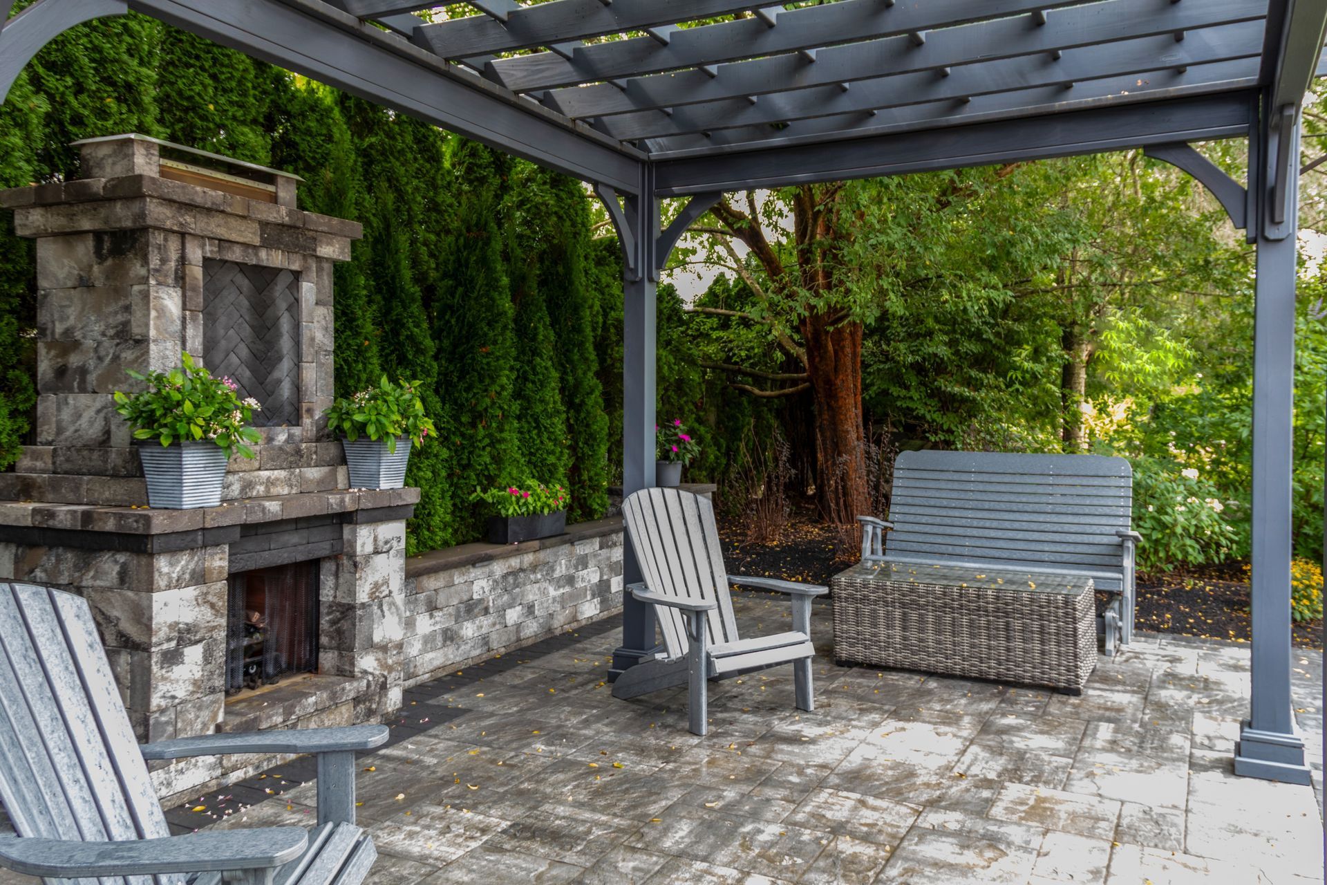 Patio with stone fireplace, gray pergola, Adirondack chairs, and bench. Lush greenery in the background.