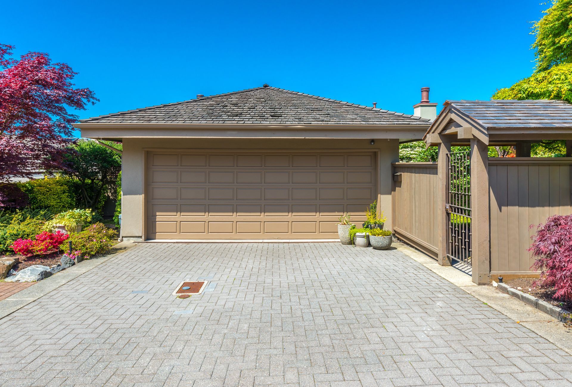 Tan garage with paved driveway, flanked by colorful plants under a bright blue sky.