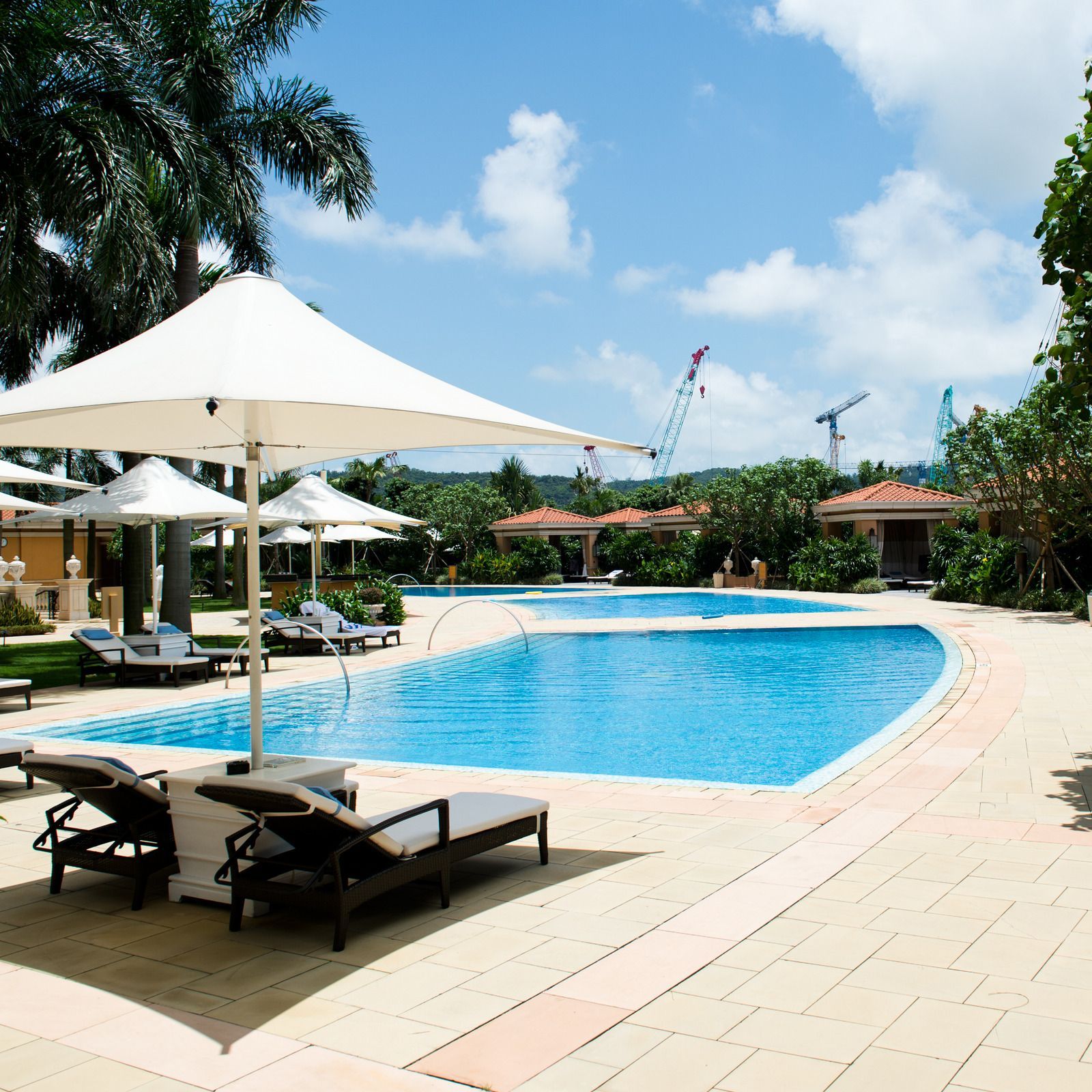 Pool area with lounge chairs under umbrellas, palm trees, and blue sky.
