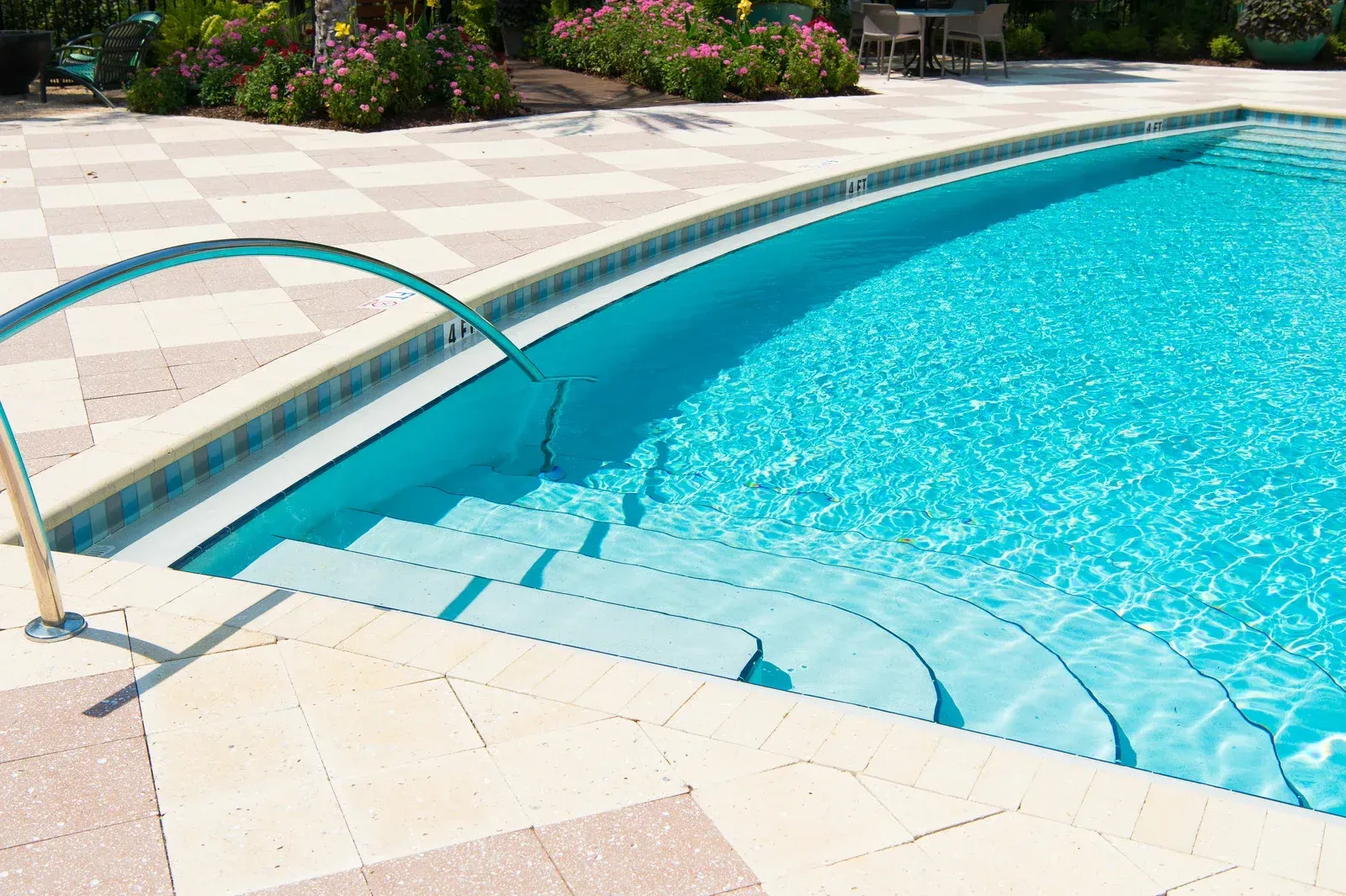 Swimming pool with curved steps, blue water, and a handrail. Paved patio, flowers, and outdoor seating in the background.