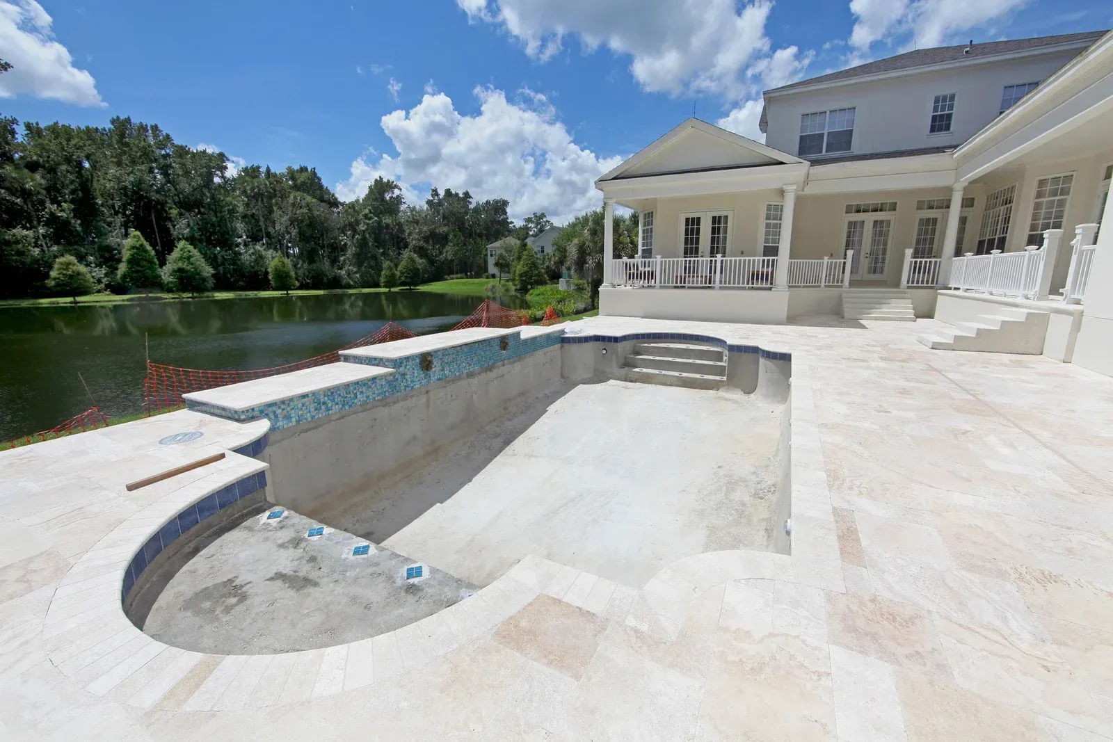 An empty pool with blue tile trim next to a white house overlooking a lake under a blue sky.