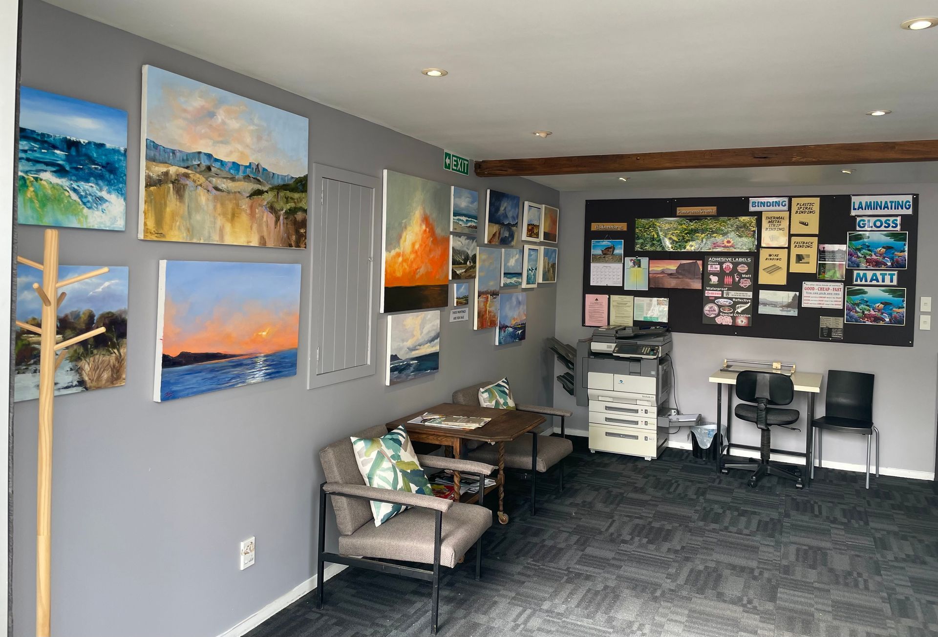 Gray-walled art studio with colorful landscape paintings, seating area, bulletin board, and copy machine.