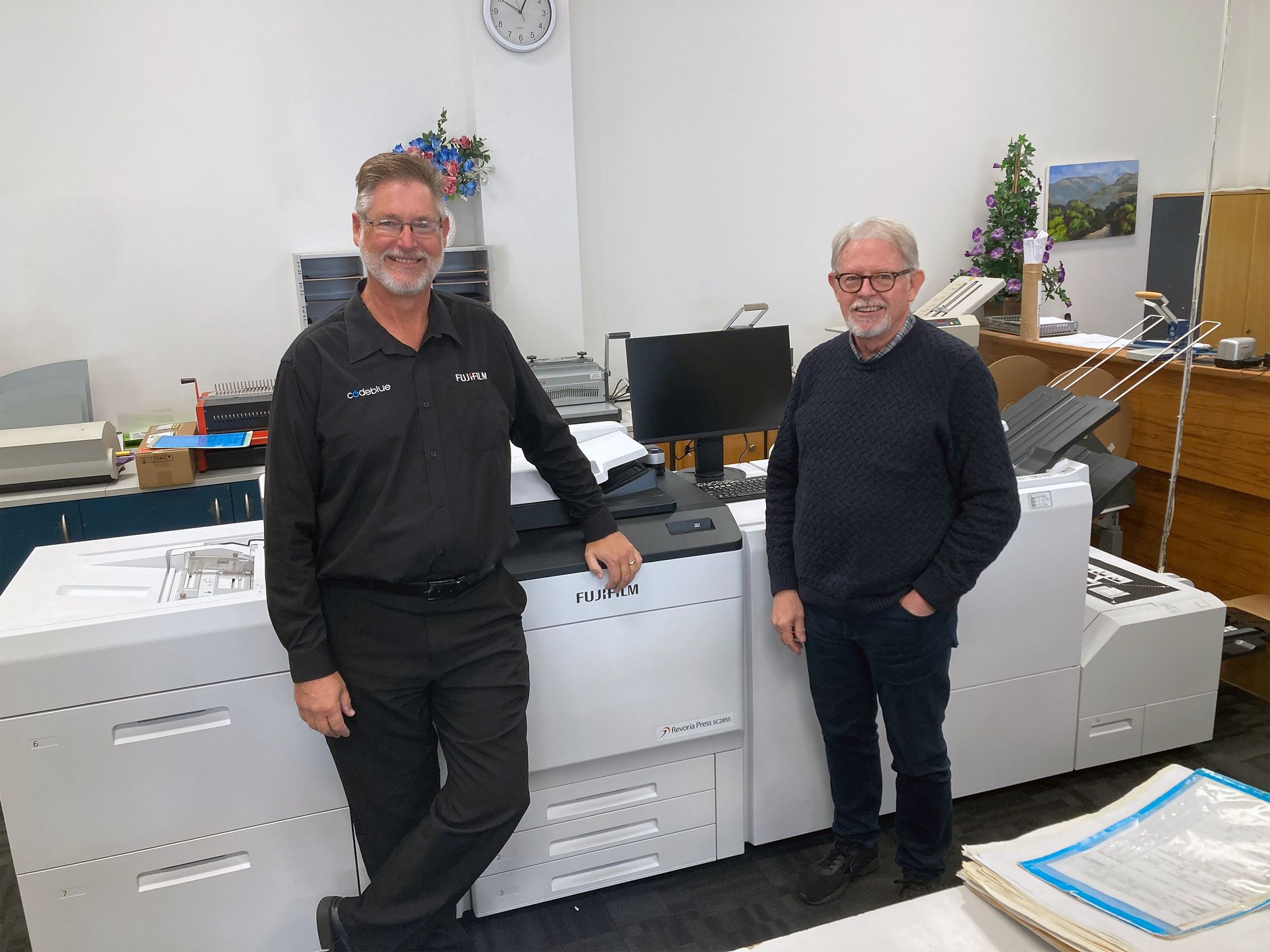 Two men standing by a large printing machine in an office. One man leans on the machine.