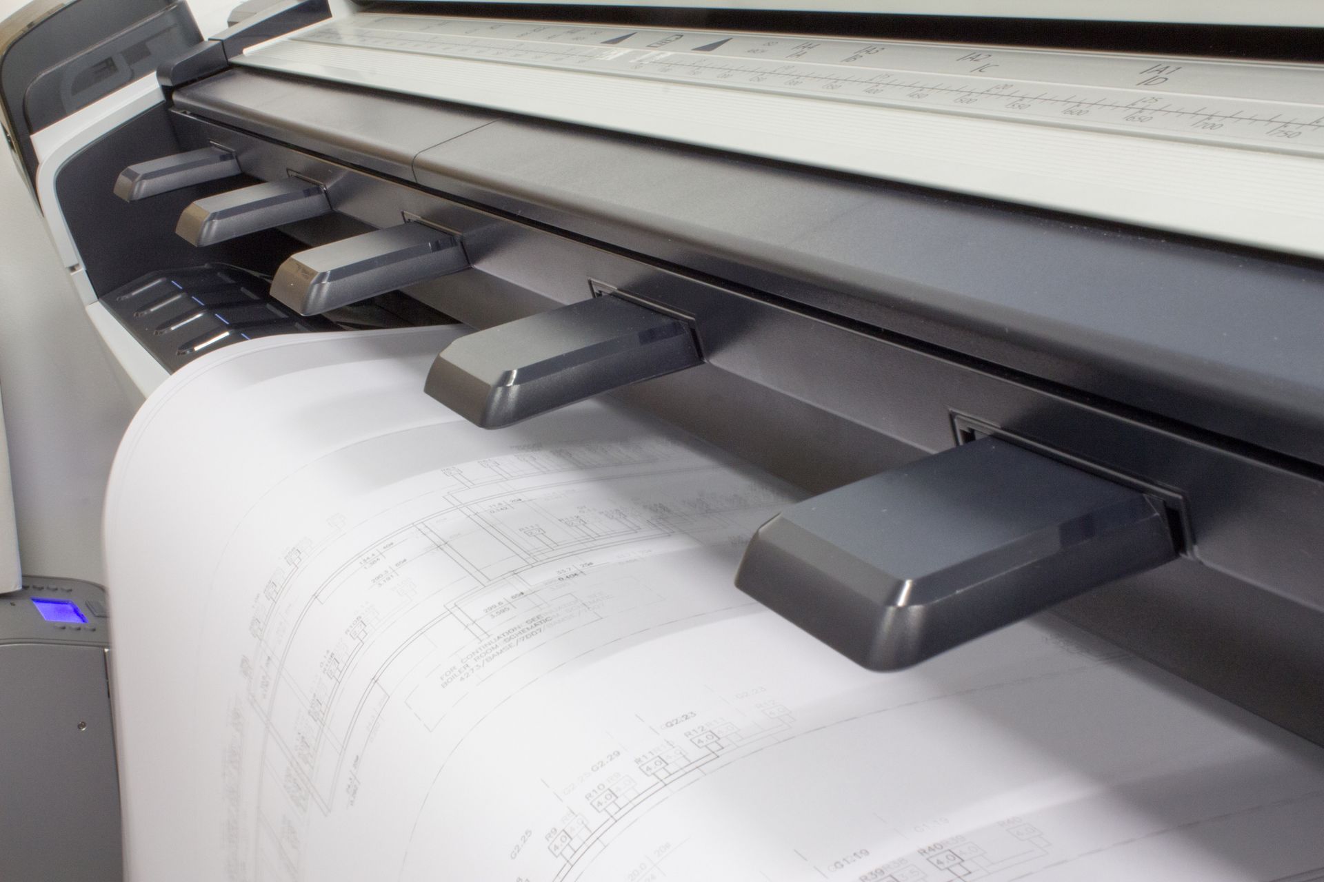 Large-format printer printing blueprints; white paper rolls out from beneath gray control panel.