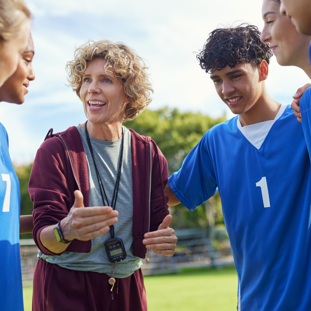A coach in maroon sweatsuit talks with a team of young people in blue jerseys on a soccer field.