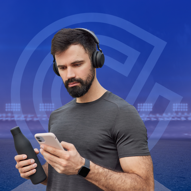 Man in headphones, looking at phone, holding water bottle, standing near stadium.