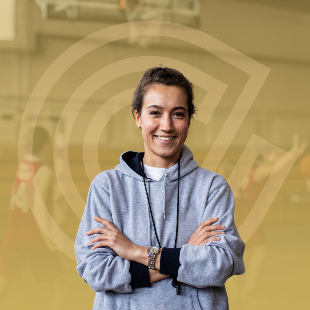 Woman in grey hoodie smiles, arms crossed, in a basketball gym.