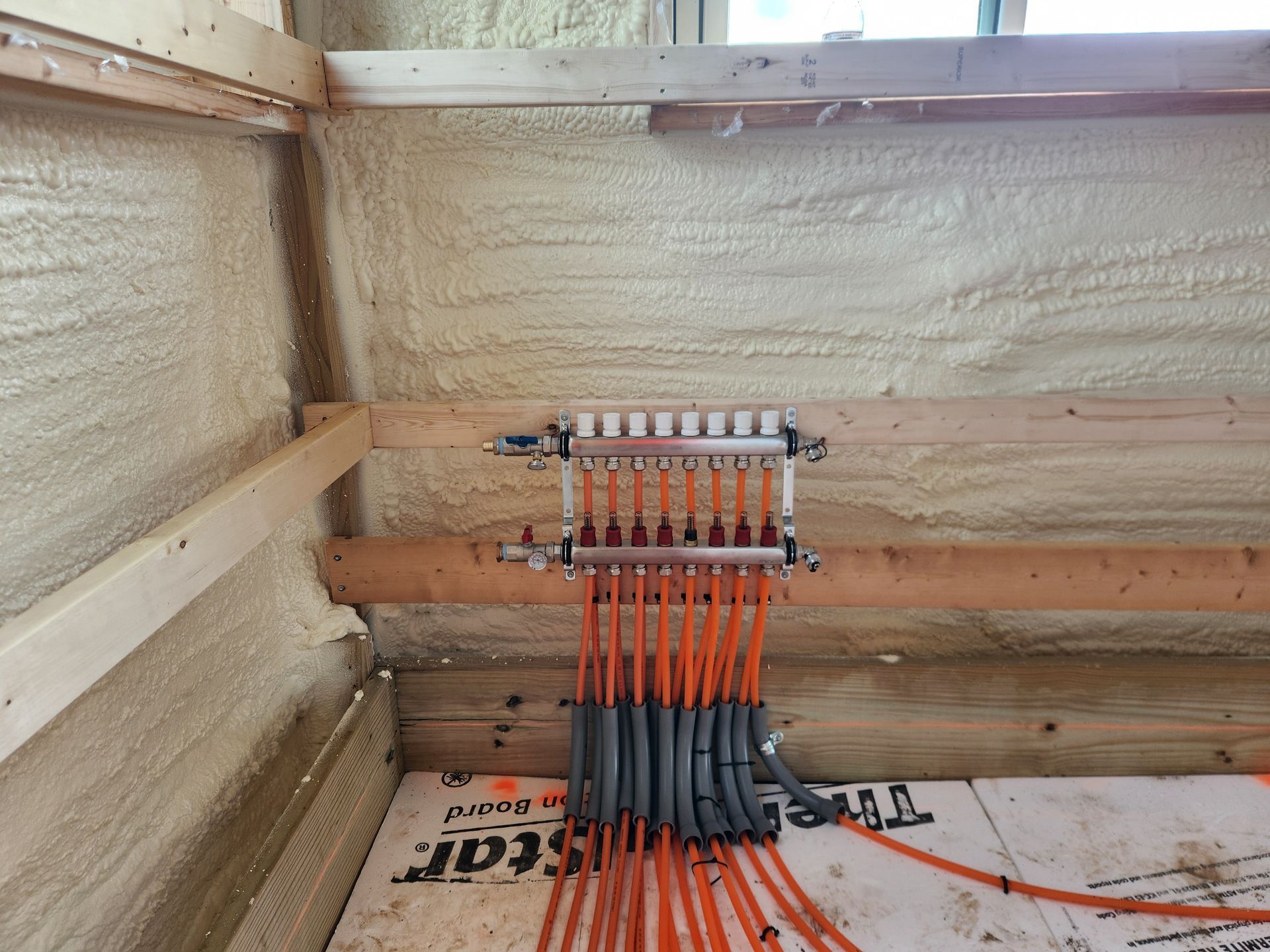 Insulated room corner with a manifold and orange tubing. Wood framing and flooring visible.