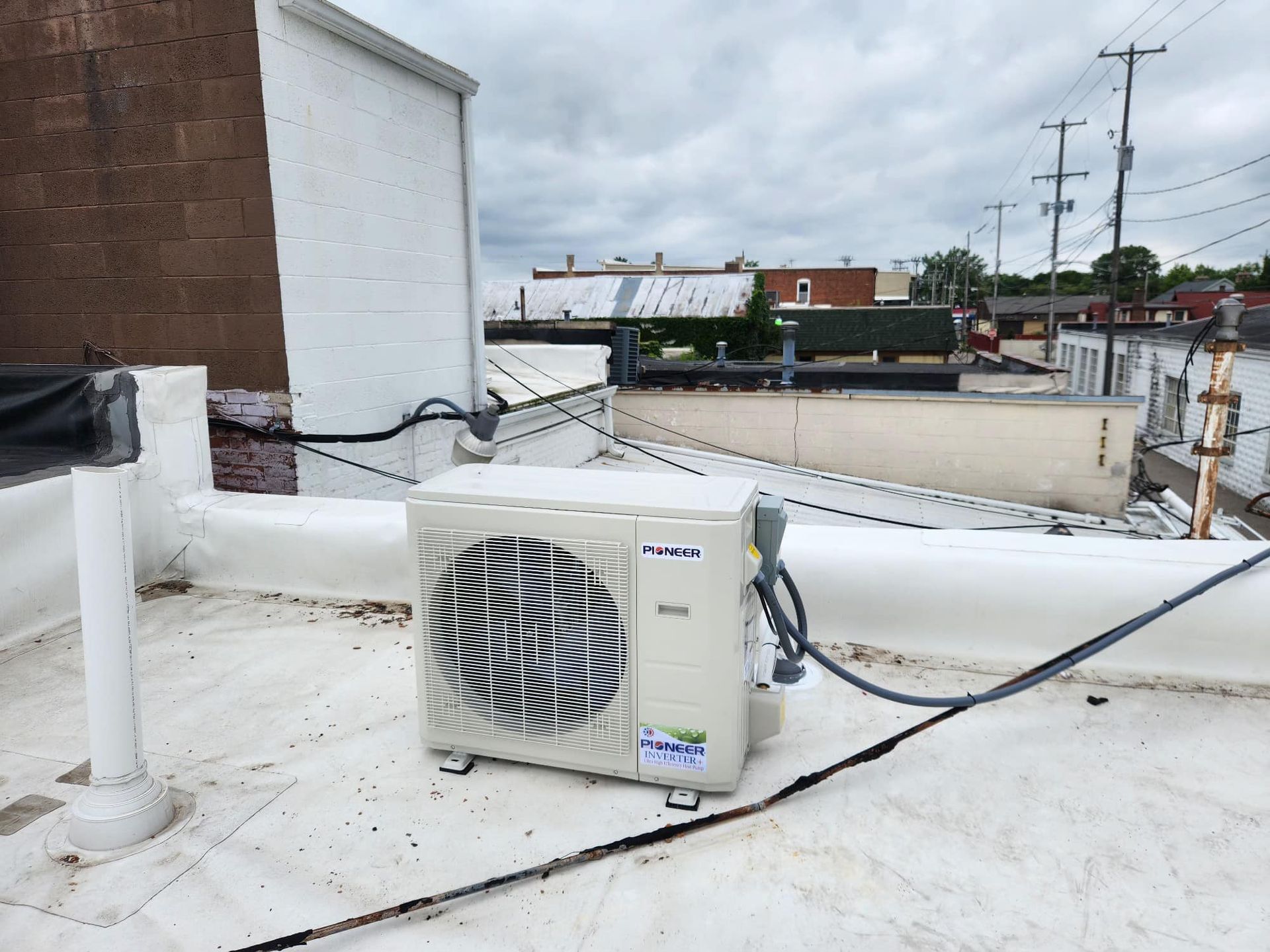 HVAC unit on a white rooftop. Dark wires and buildings in the background under a cloudy sky.