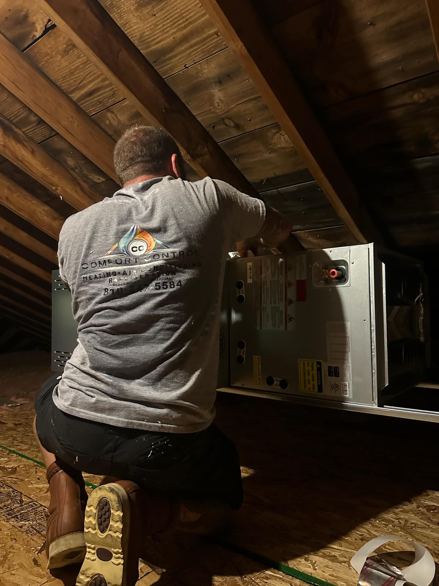 HVAC technician in an attic installing equipment. He wears a gray t-shirt and kneels near ductwork.
