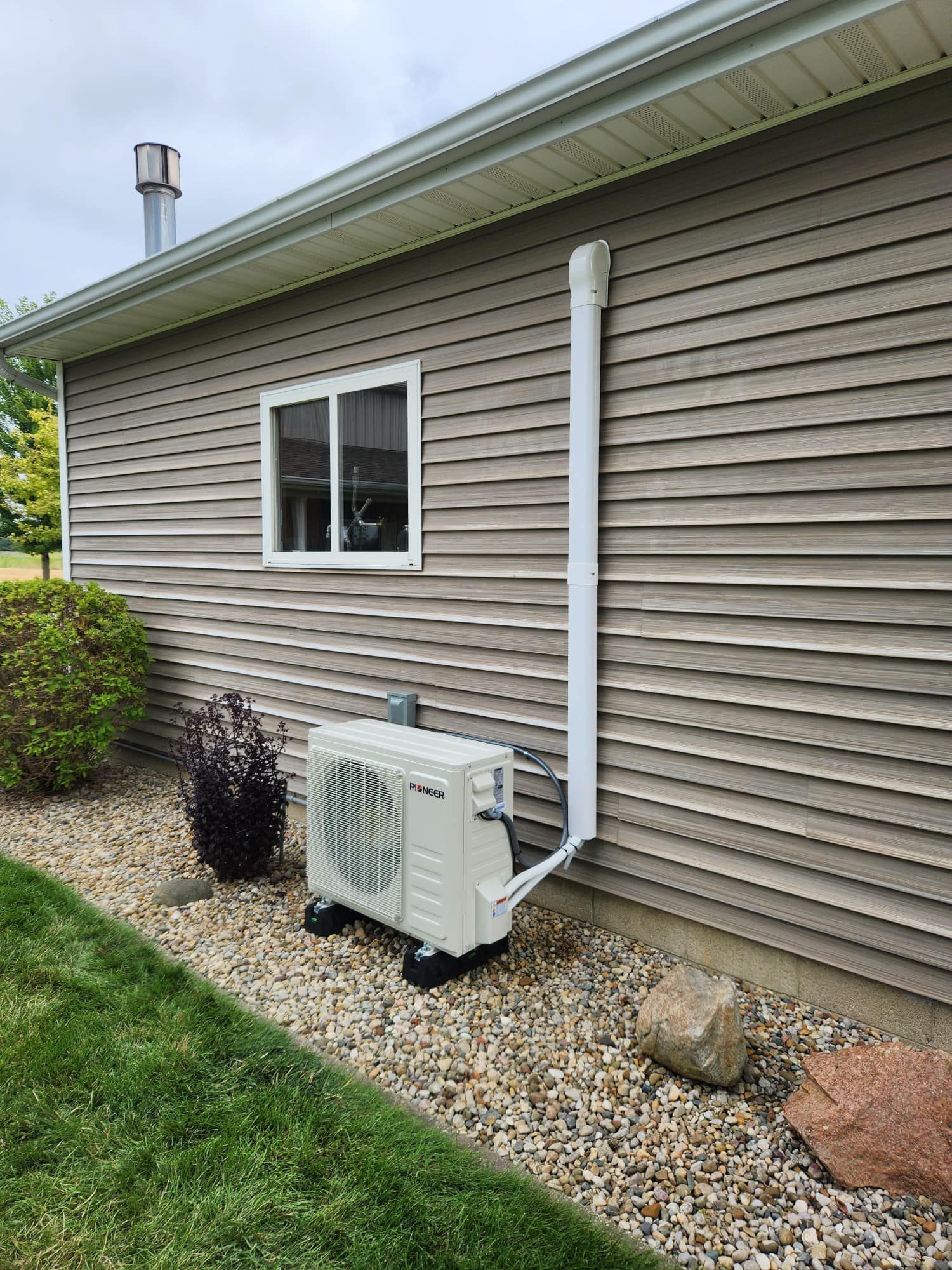 Air conditioner unit on gravel beside a house with tan siding, next to a window and white pipe.
