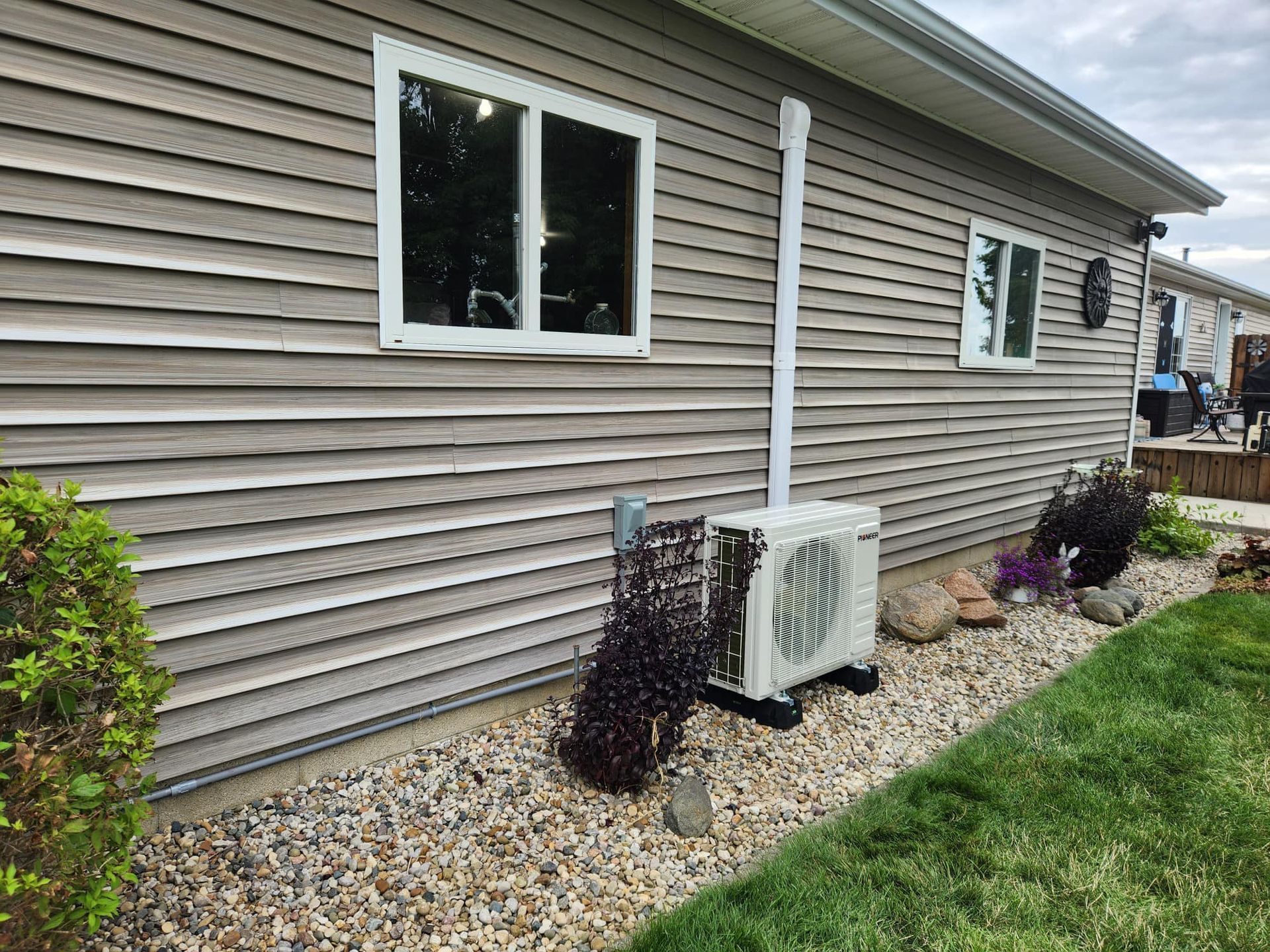 An air conditioning unit sits outside a house with siding and windows, beside landscaping stones and grass.