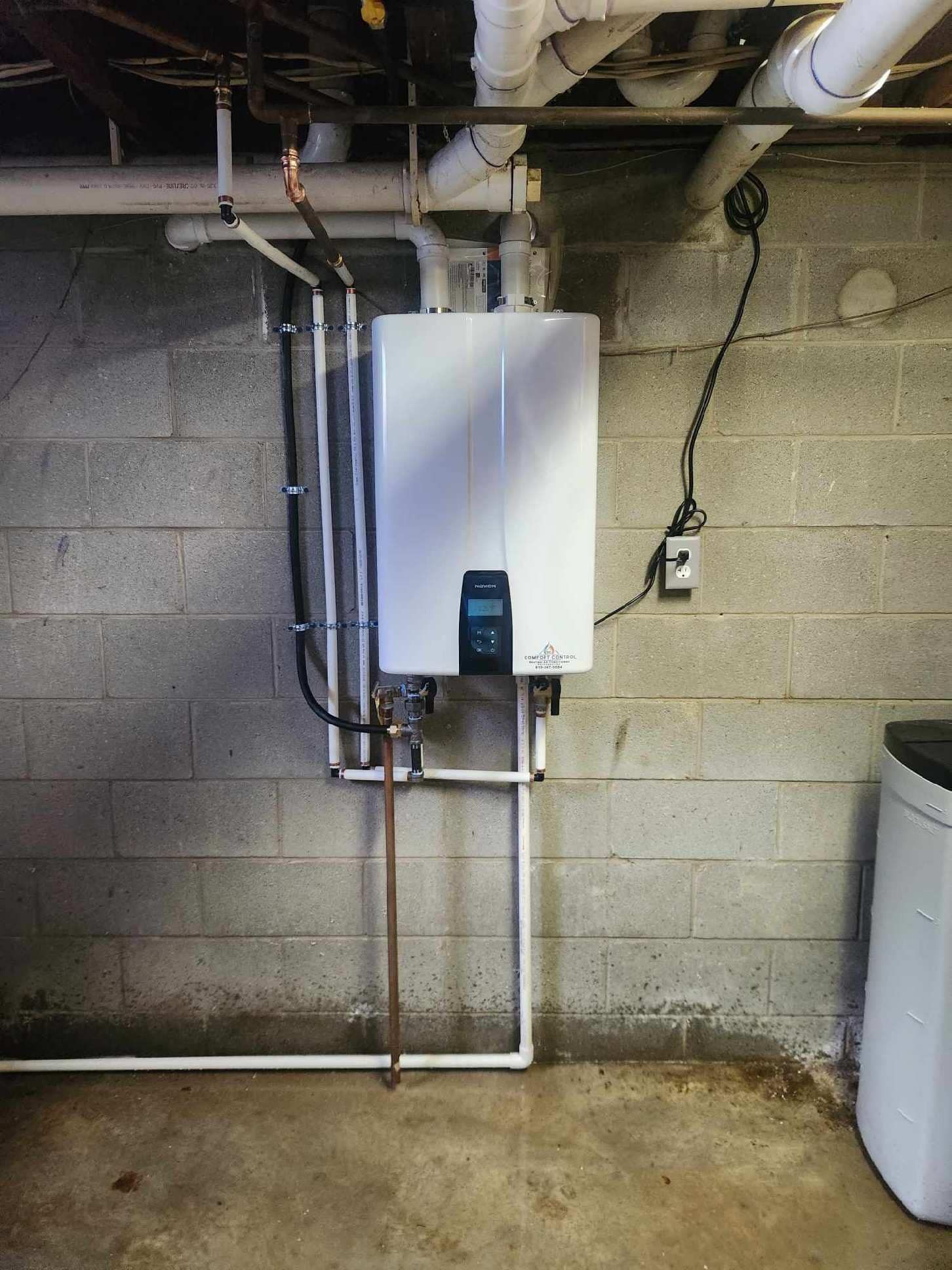 White tankless water heater mounted on a gray cinder block wall. Pipes and wires are connected. Concrete floor.