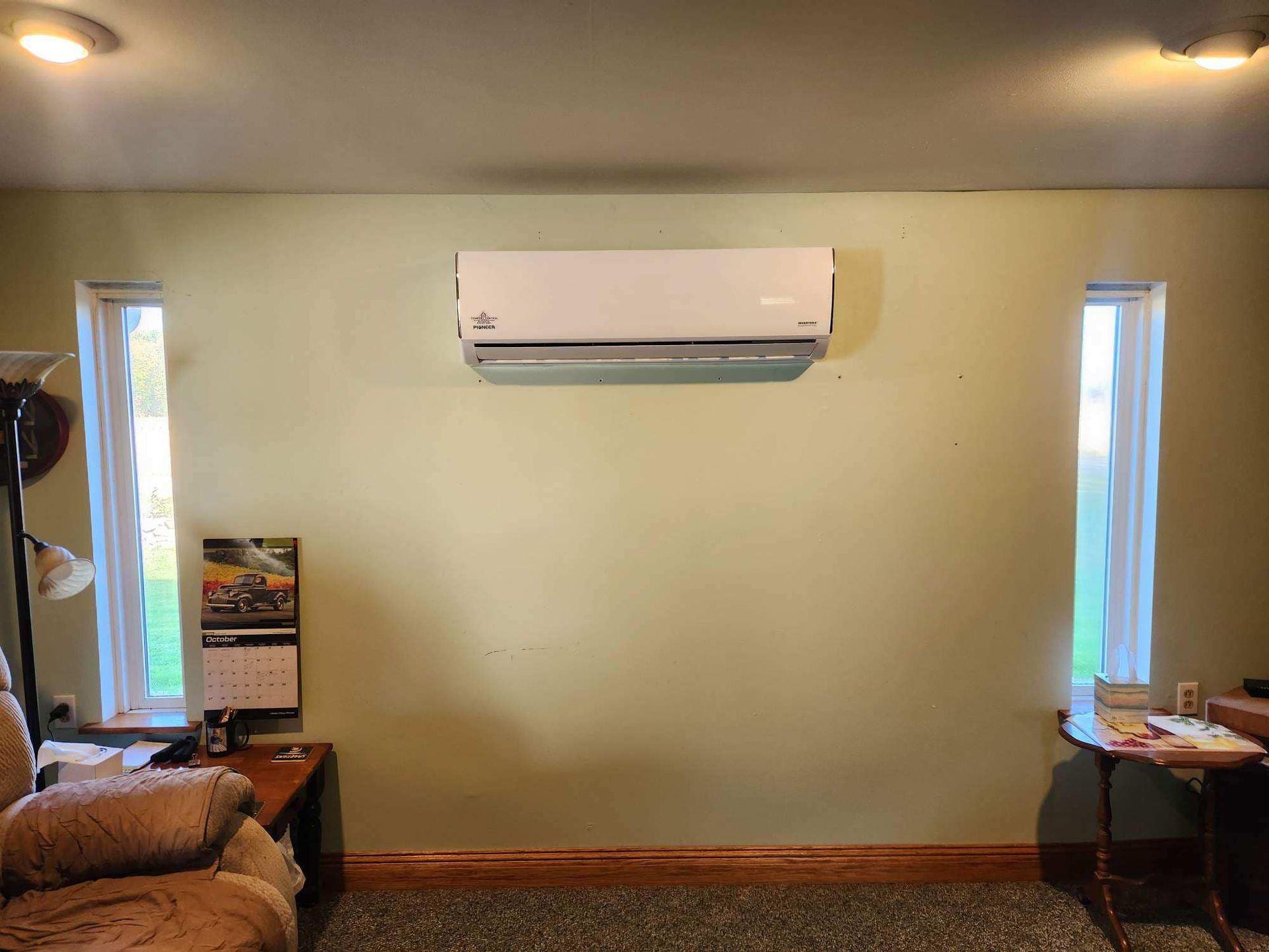 Wall-mounted air conditioner centered above a wood baseboard, flanked by tall narrow windows on a green wall.