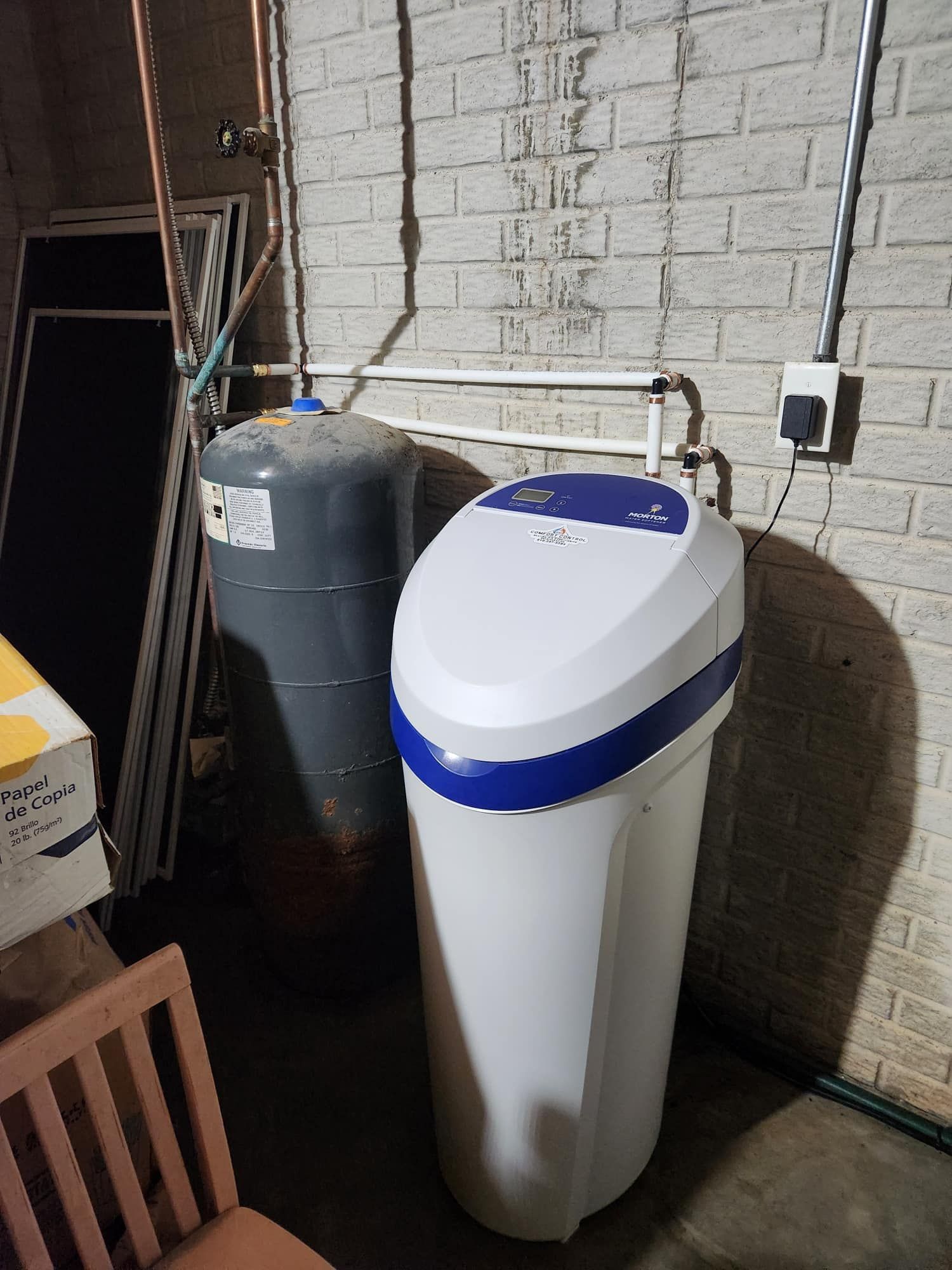 Water softener and pressure tank against a brick wall in a basement. White and blue softener, grey tank.