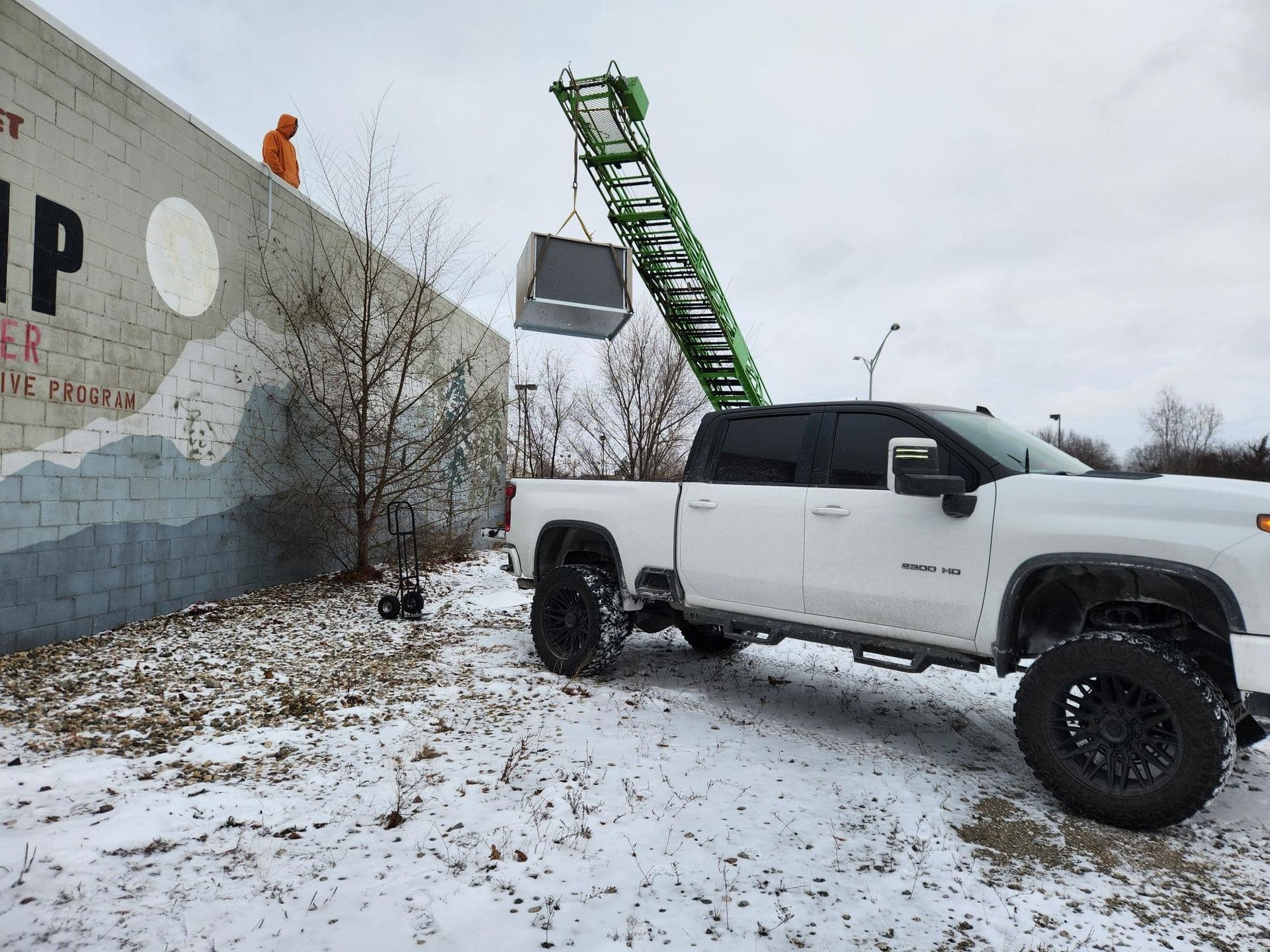 A white pickup truck uses a lift to move a large, dark object over a painted wall in a snowy setting.