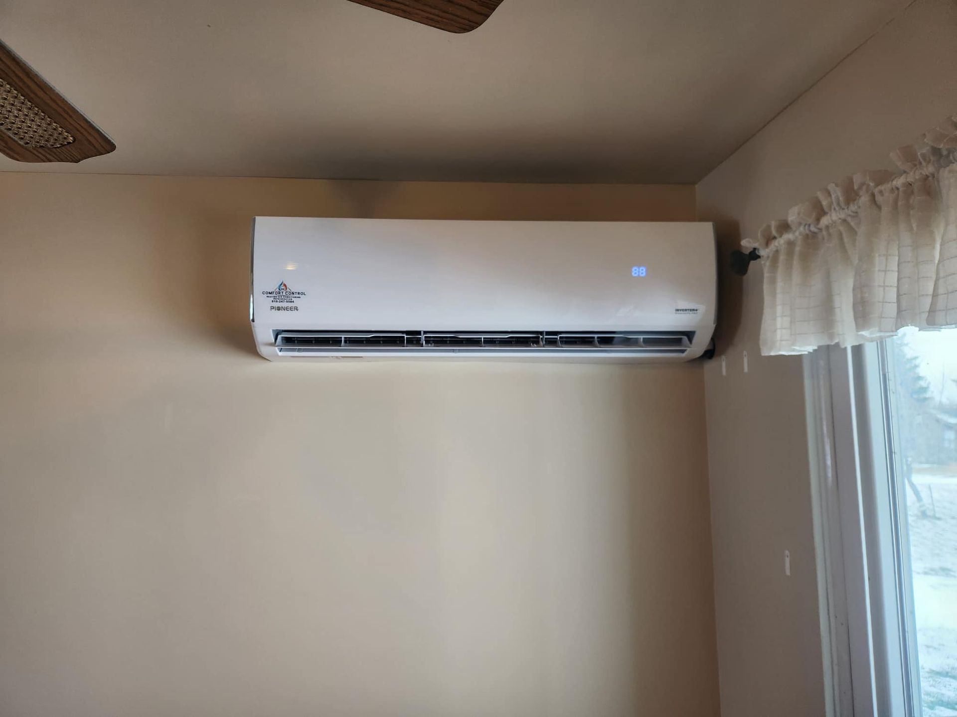 White wall-mounted air conditioner unit above a wall vent. Tan wall and window with white trim in the background.