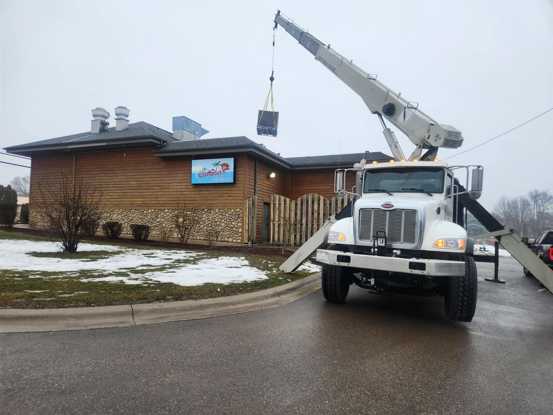 A crane lifting an object over a one-story building with a log-cabin-style exterior on a cloudy day.