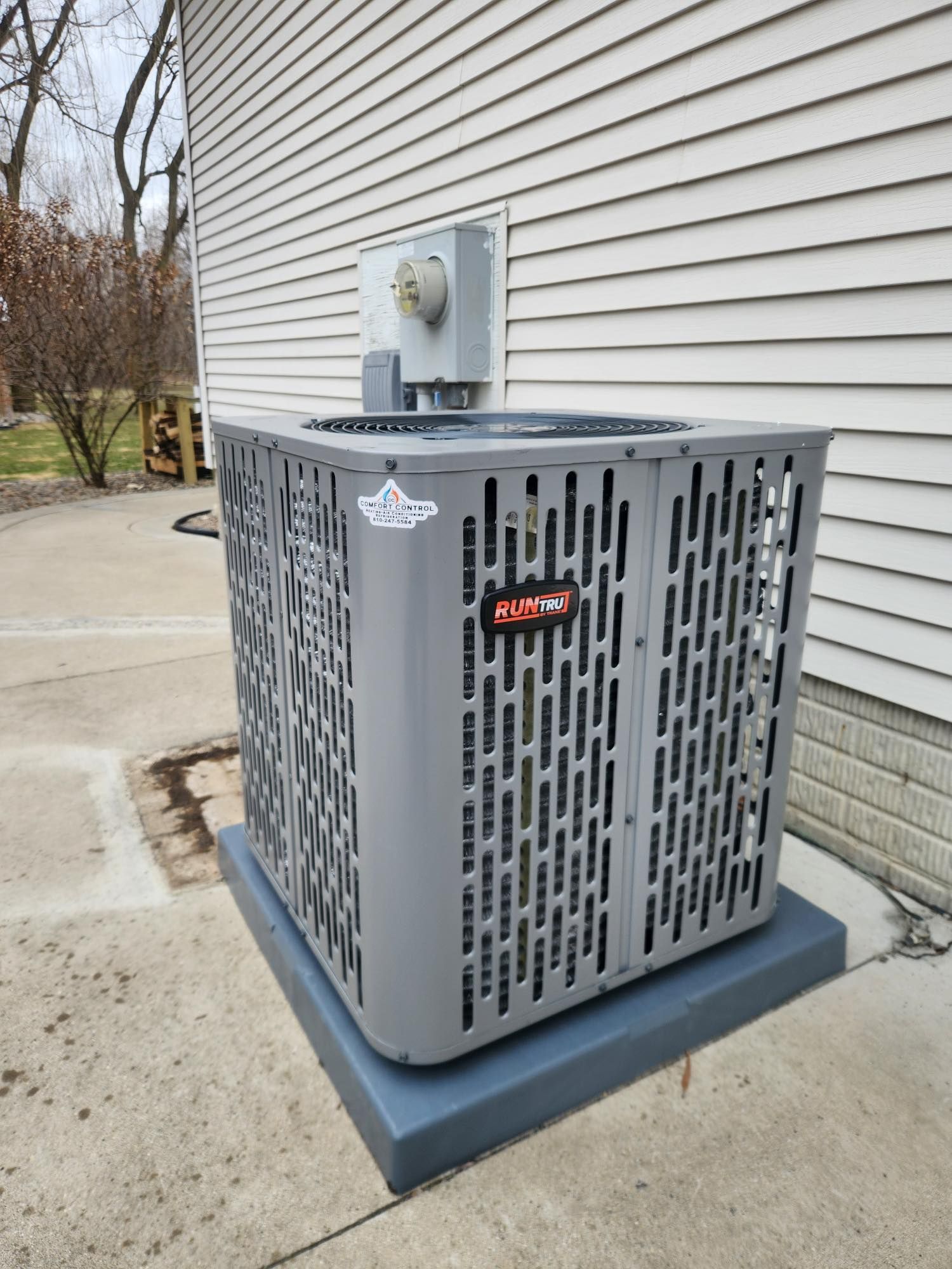 Gray air conditioning unit on a concrete pad next to a beige house, with electrical box above.