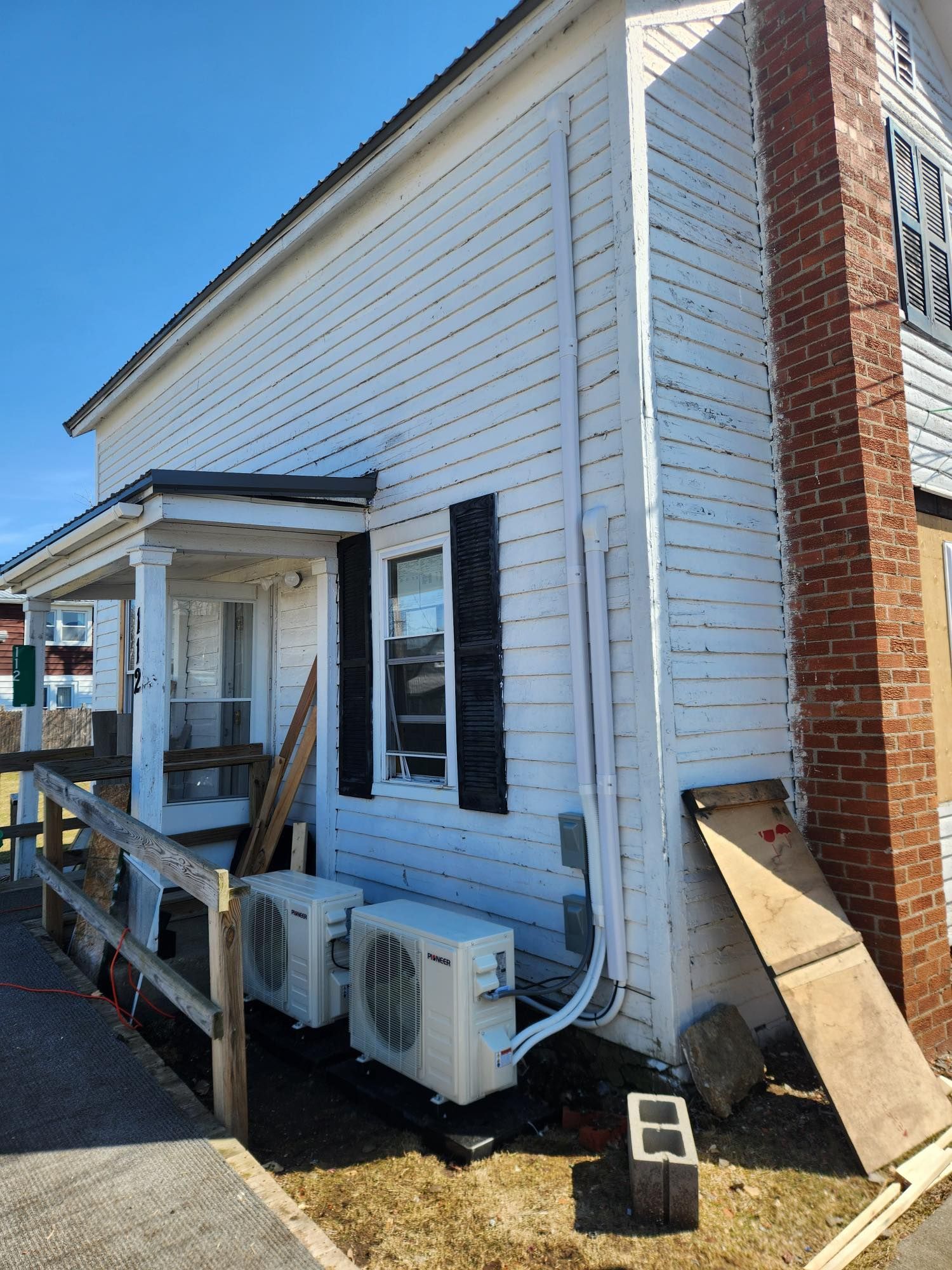 White building with porch, window, black shutters, brick chimney, and air conditioning units.