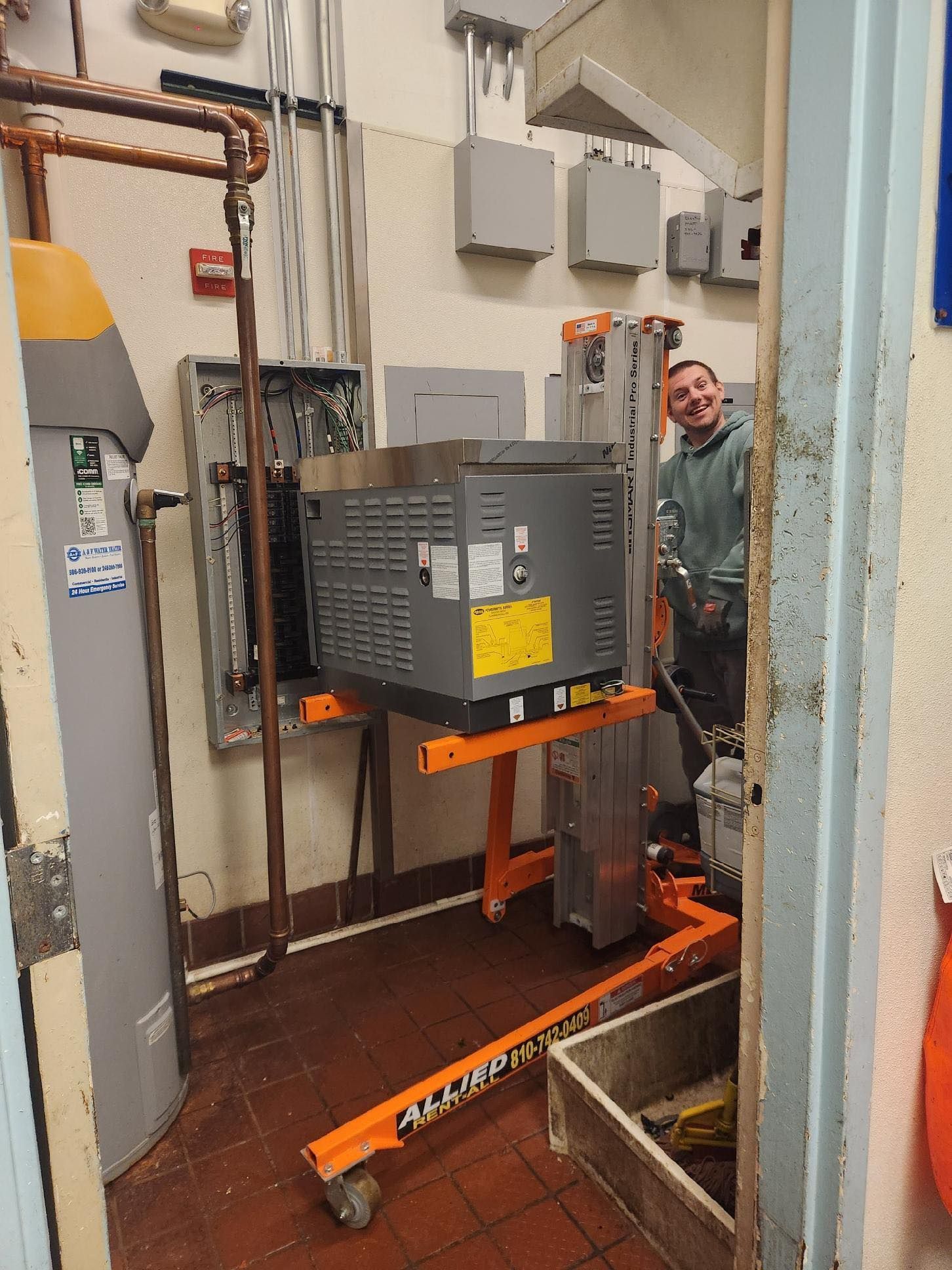 Man using lift to move large appliance in a utility room; water heater, electrical panel visible.