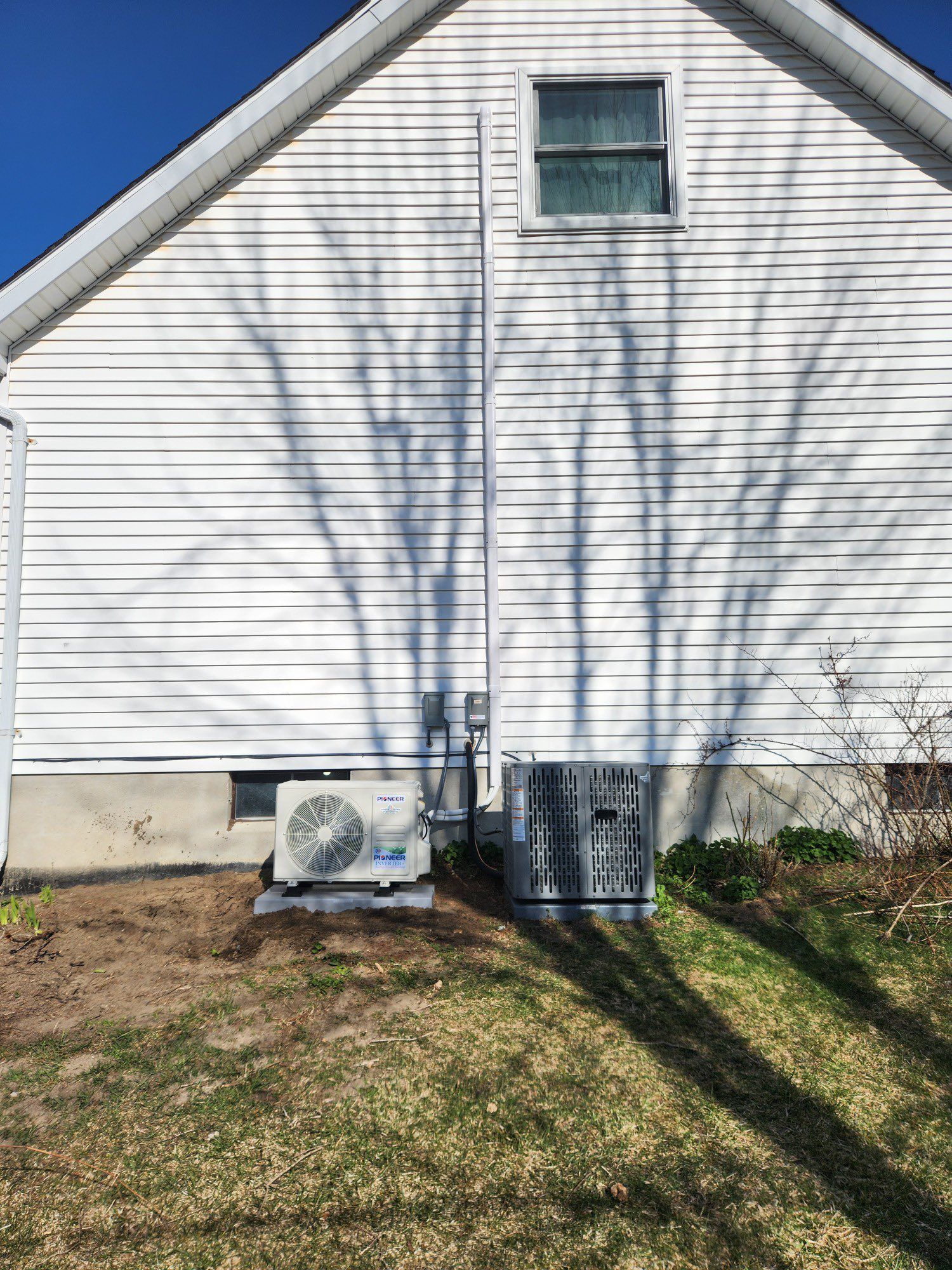 Two air conditioning units sit beside a white house, with a tree shadow cast on the wall.