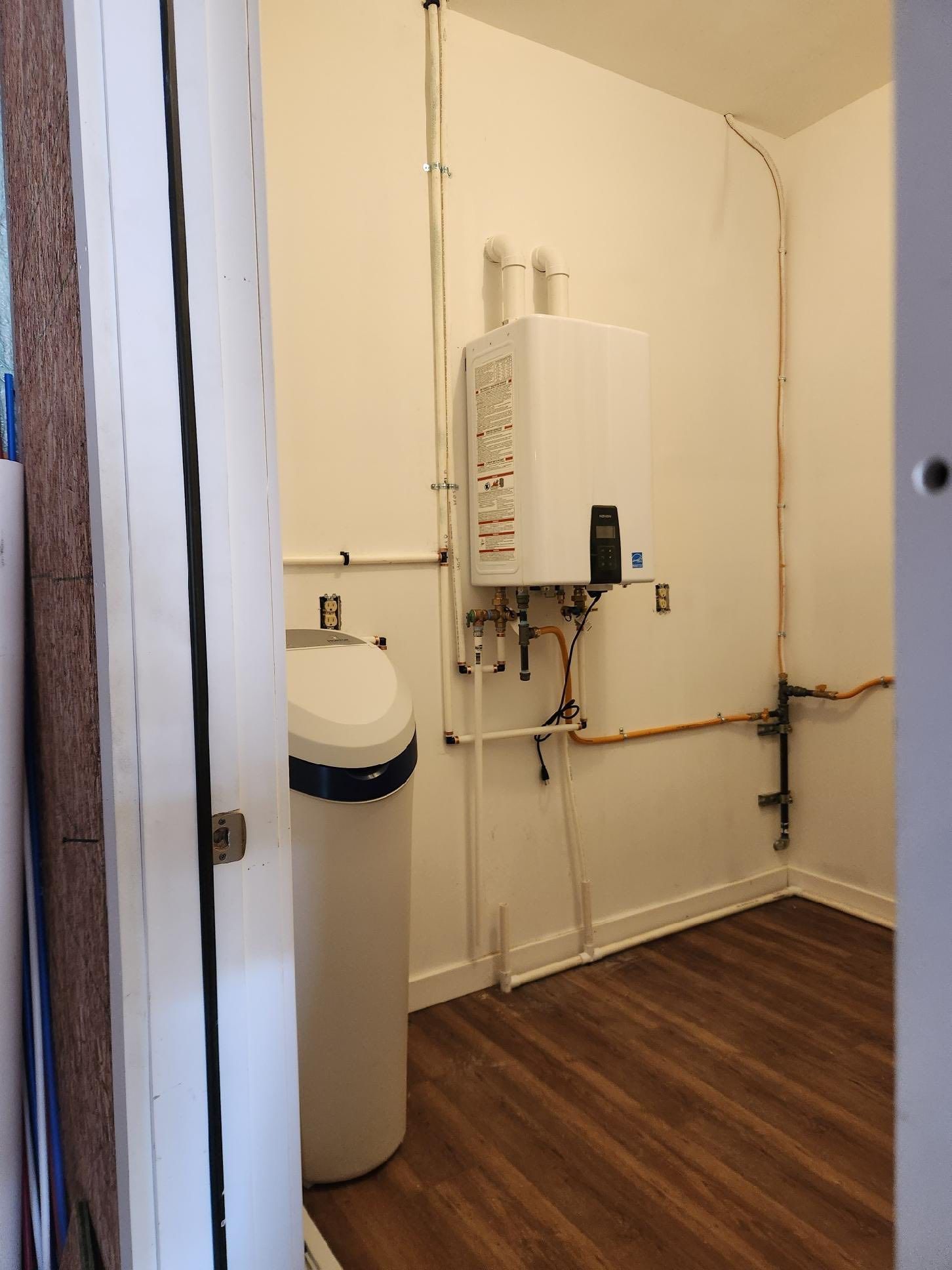Utility room with tankless water heater, water softener, and pipes on white walls. Brown floor.
