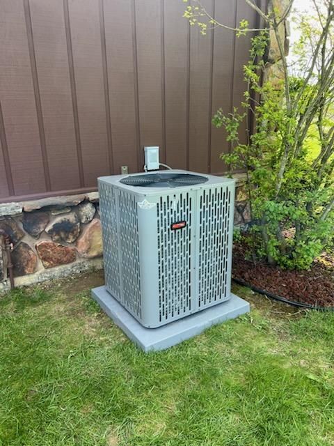 Gray air conditioning unit on a concrete base, next to a brown wall and green bushes.