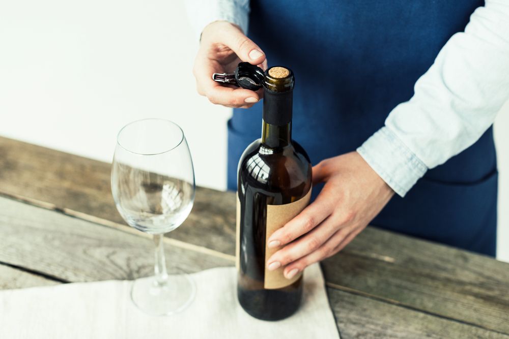 Persona che apre una bottiglia di vino con un cavatappi vicino a un bicchiere di vino su un tavolo di legno.