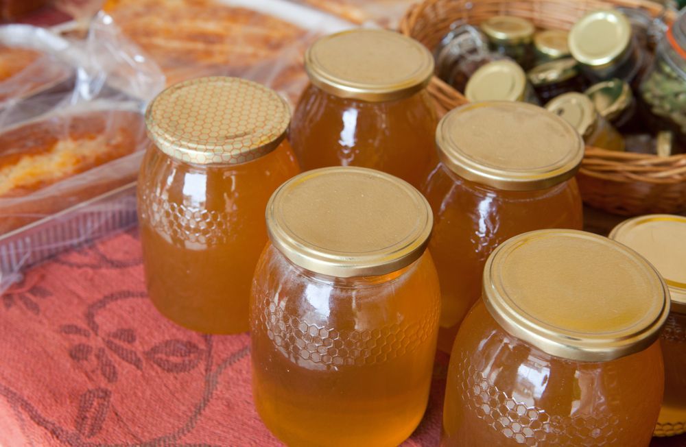 Barattoli di miele dorato con coperchi dorati, appoggiati su un panno rosso, con prodotti da forno e barattoli sullo sfondo.