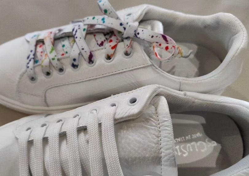 Un paio di scarpe da ginnastica bianche: una con lacci colorati e macchiati di vernice e l'altra con lacci bianchi semplici.