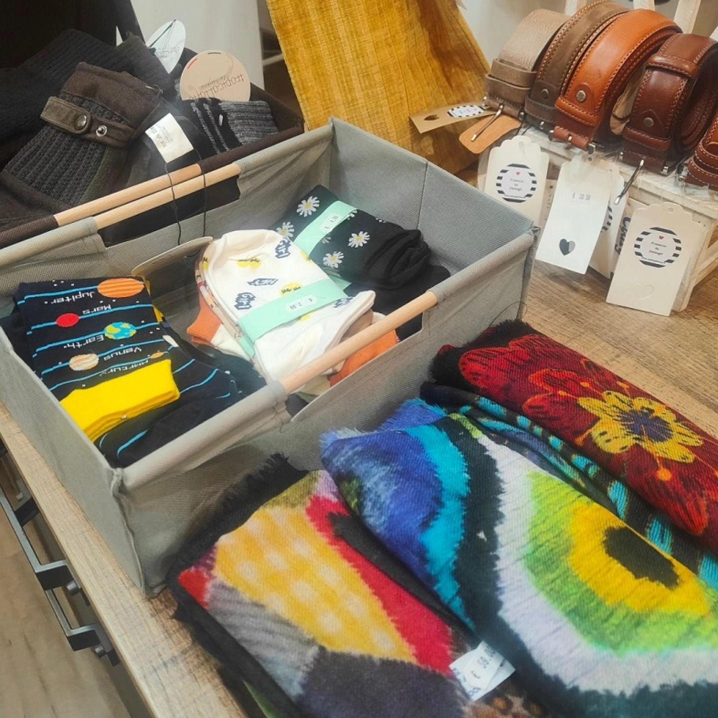 In un contenitore sono esposti calzini piegati e sciarpe colorate a fantasia, vicino sono appese cinture di pelle.