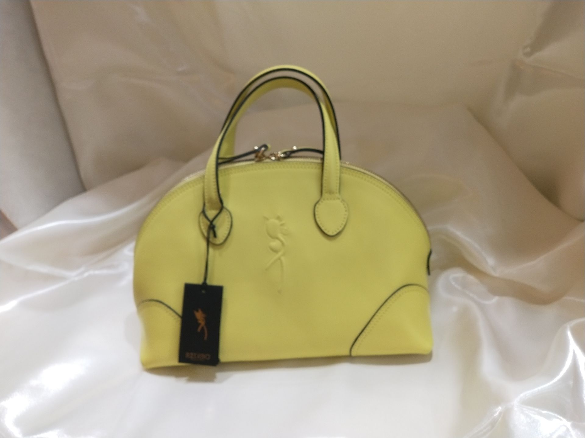 Una borsa a mano giallo brillante, a forma di cupola, con manici superiori e un'etichetta con logo, sfondo di tessuto bianco.