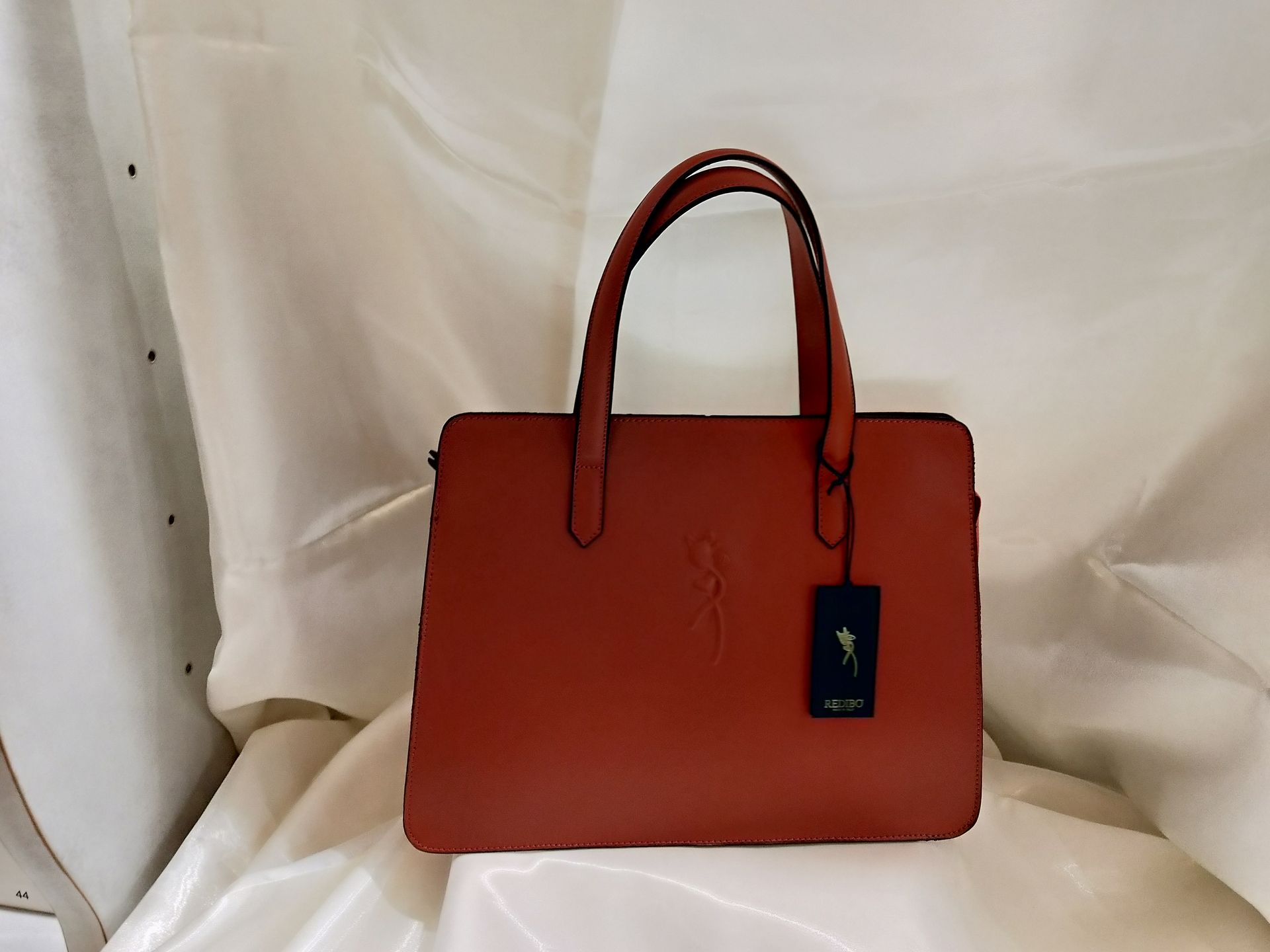 Borsa strutturata color arancio ruggine con due manici superiori e un'etichetta rettangolare con logo pendente, su uno sfondo di tessuto drappeggiato bianco.