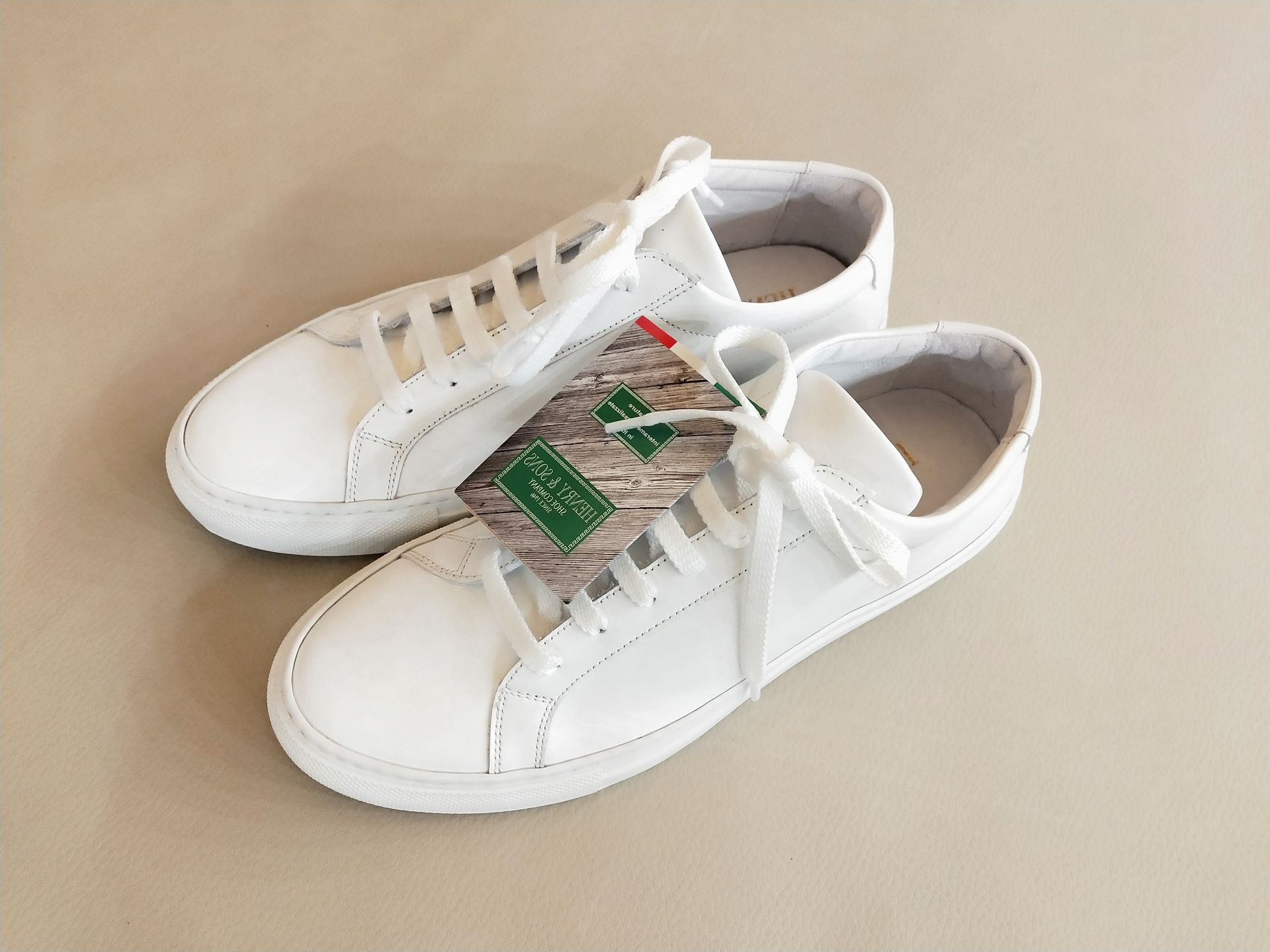 Un paio di scarpe da ginnastica in pelle bianca con lacci abbinati e un'etichetta verde del marchio, su uno sfondo beige uniforme.
