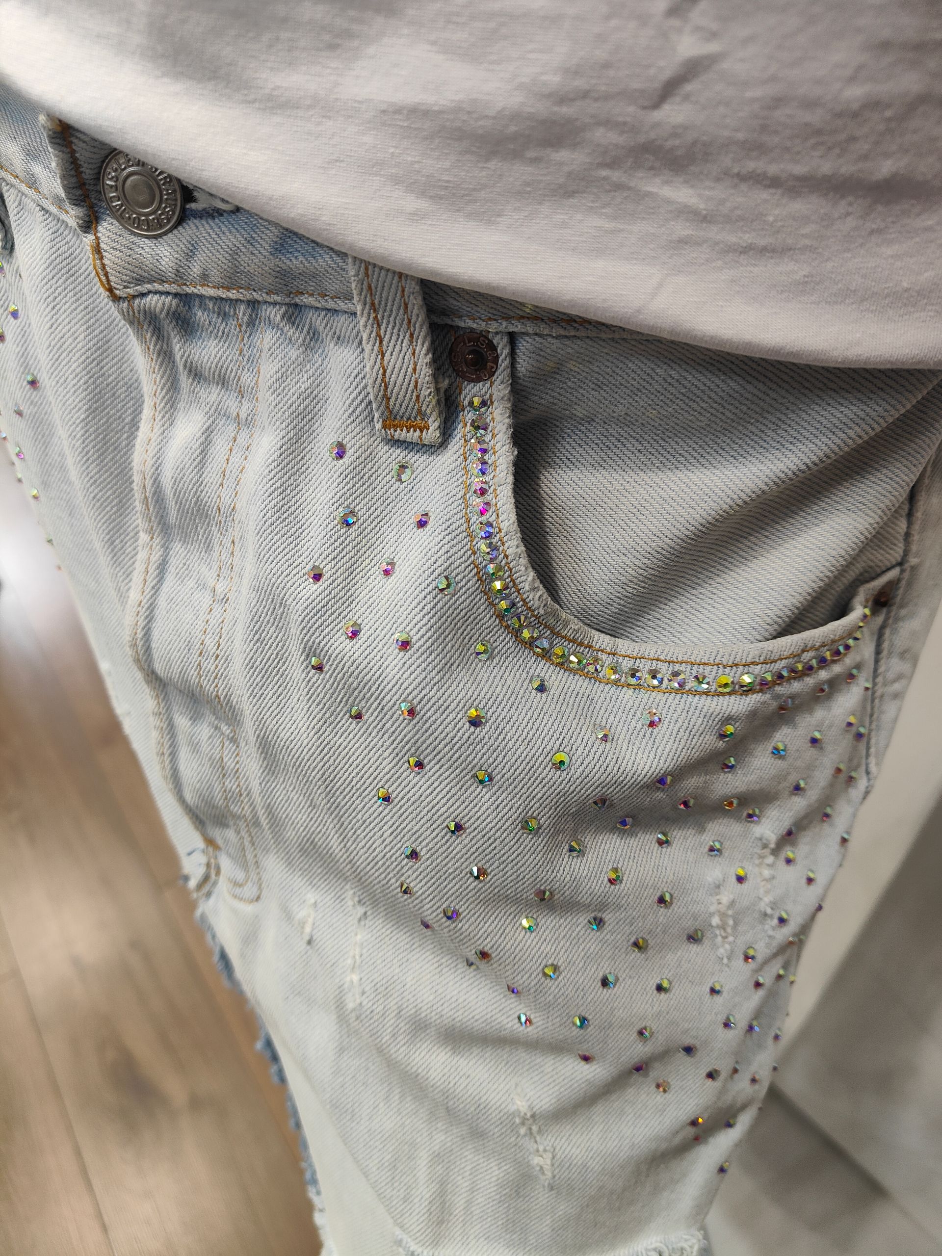 Primo piano di un paio di jeans in denim chiaro, decorati con piccoli strass multicolori intorno alla tasca frontale.